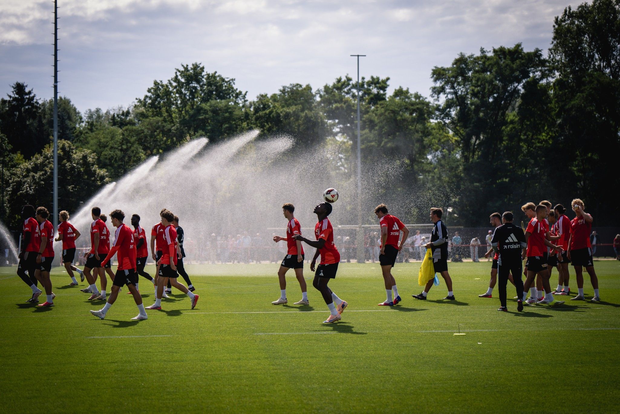 Fußballmannschaft im Training auf einem Rasenplatz, umgeben von Sprinklern und Bäumen. Spieler in roten Trikots und Trainer beobachten.