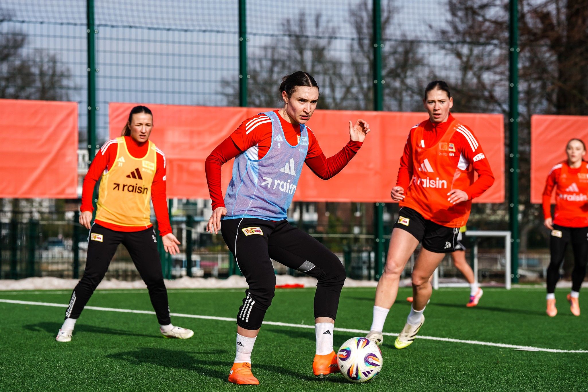 Drei weibliche Fußballspielerinnen trainieren auf einem Kunstrasenplatz, eine hat den Ball am Fuß.