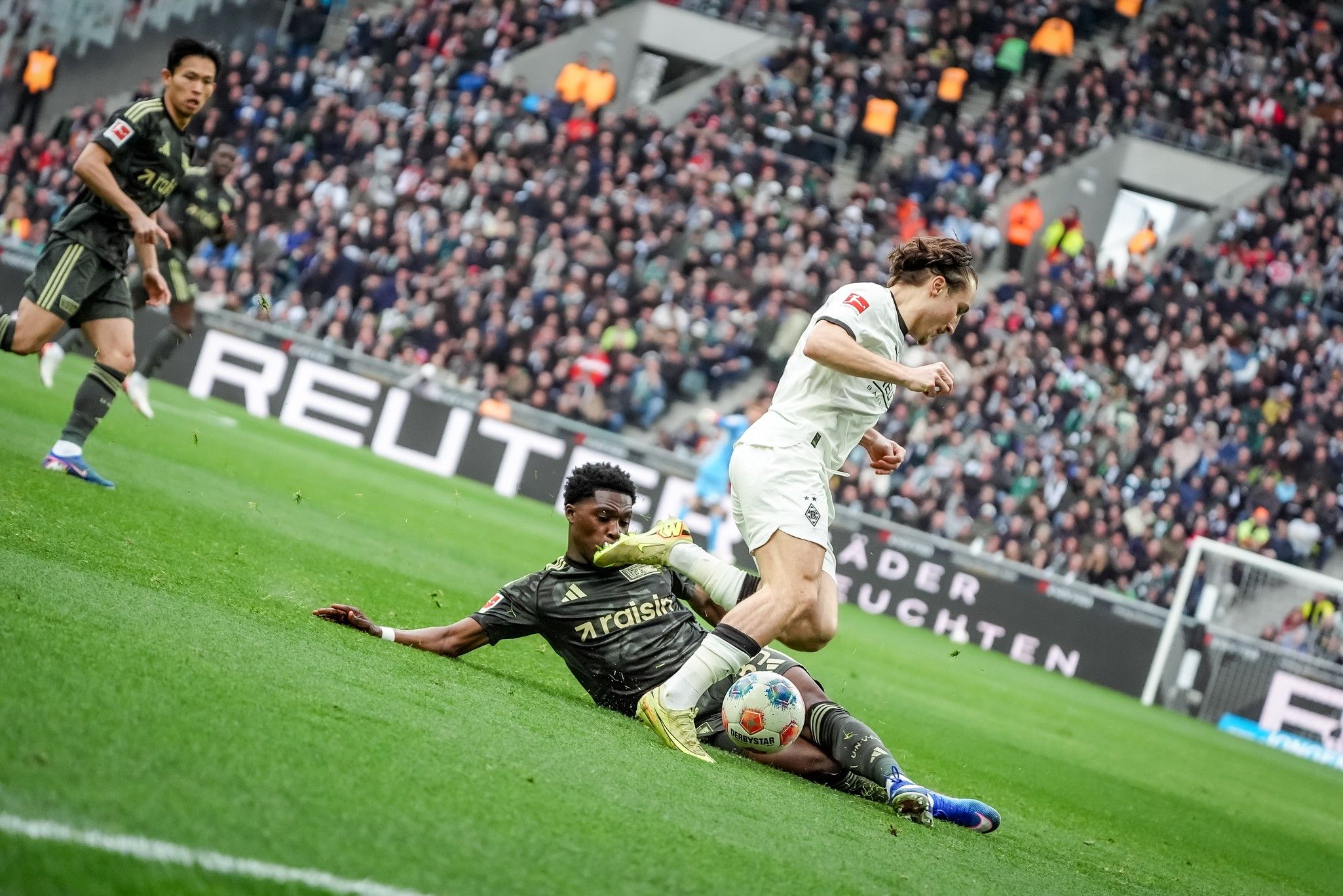 Ein Spieler in weißem Trikot weicht einem tacklenden Spieler in schwarzem Trikot aus, während ein weiterer Spieler im Hintergrund beobachtet.