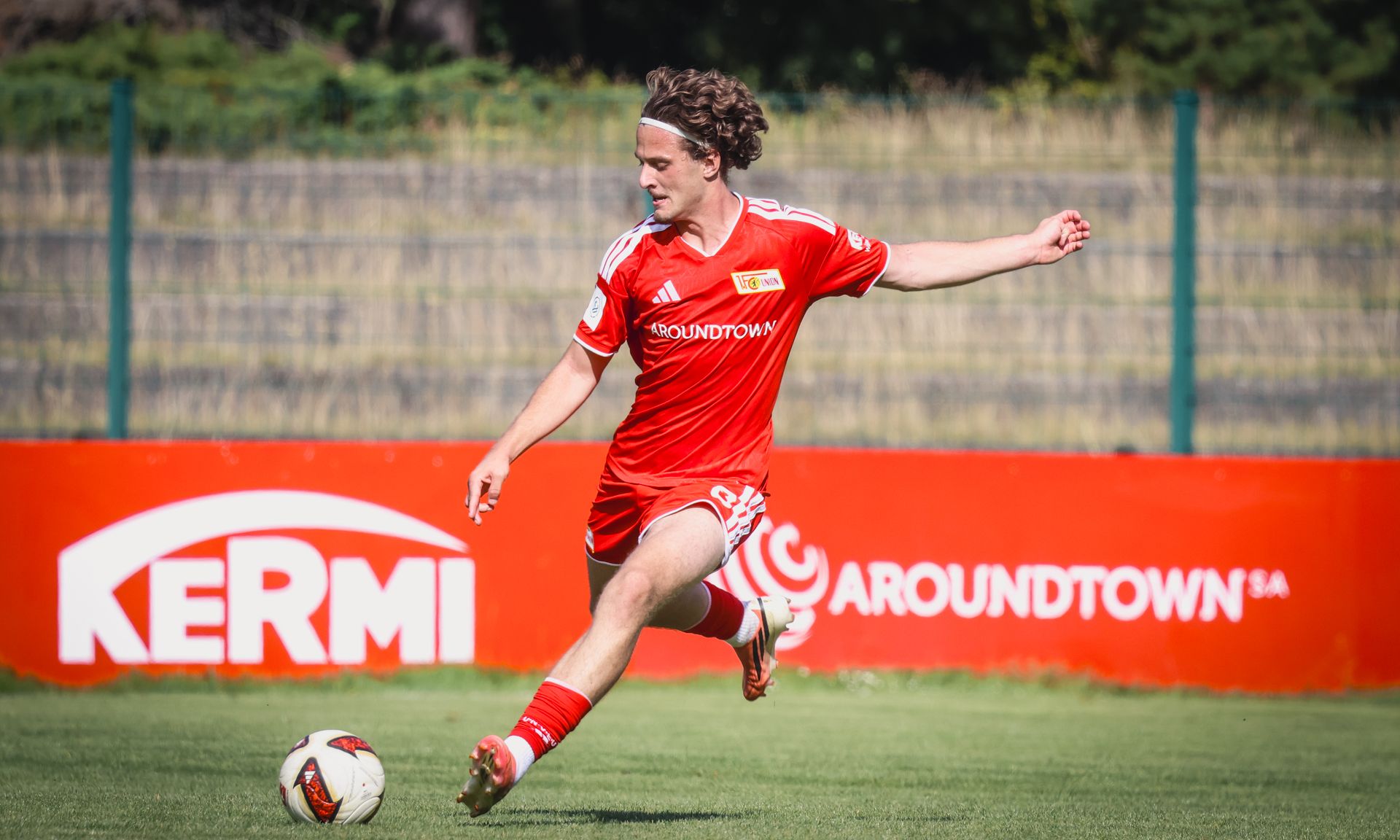 Ein Fußballspieler in rotem Trikot tritt den Ball auf einem grünen Platz vor einer Werbetafel.