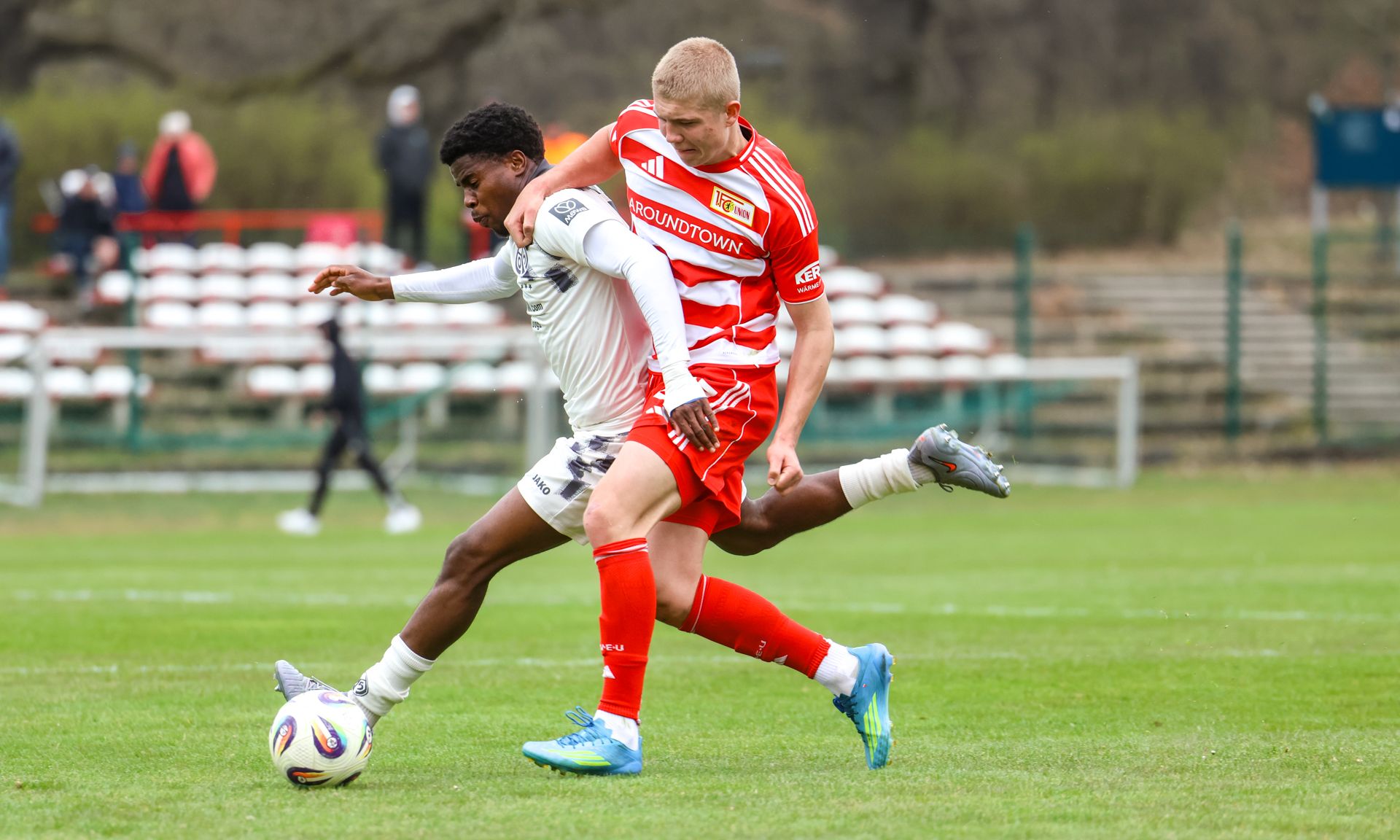 Zwei Fußballspieler kämpfen um den Ball auf einem Rasenfeld, während Zuschauer im Hintergrund stehen.