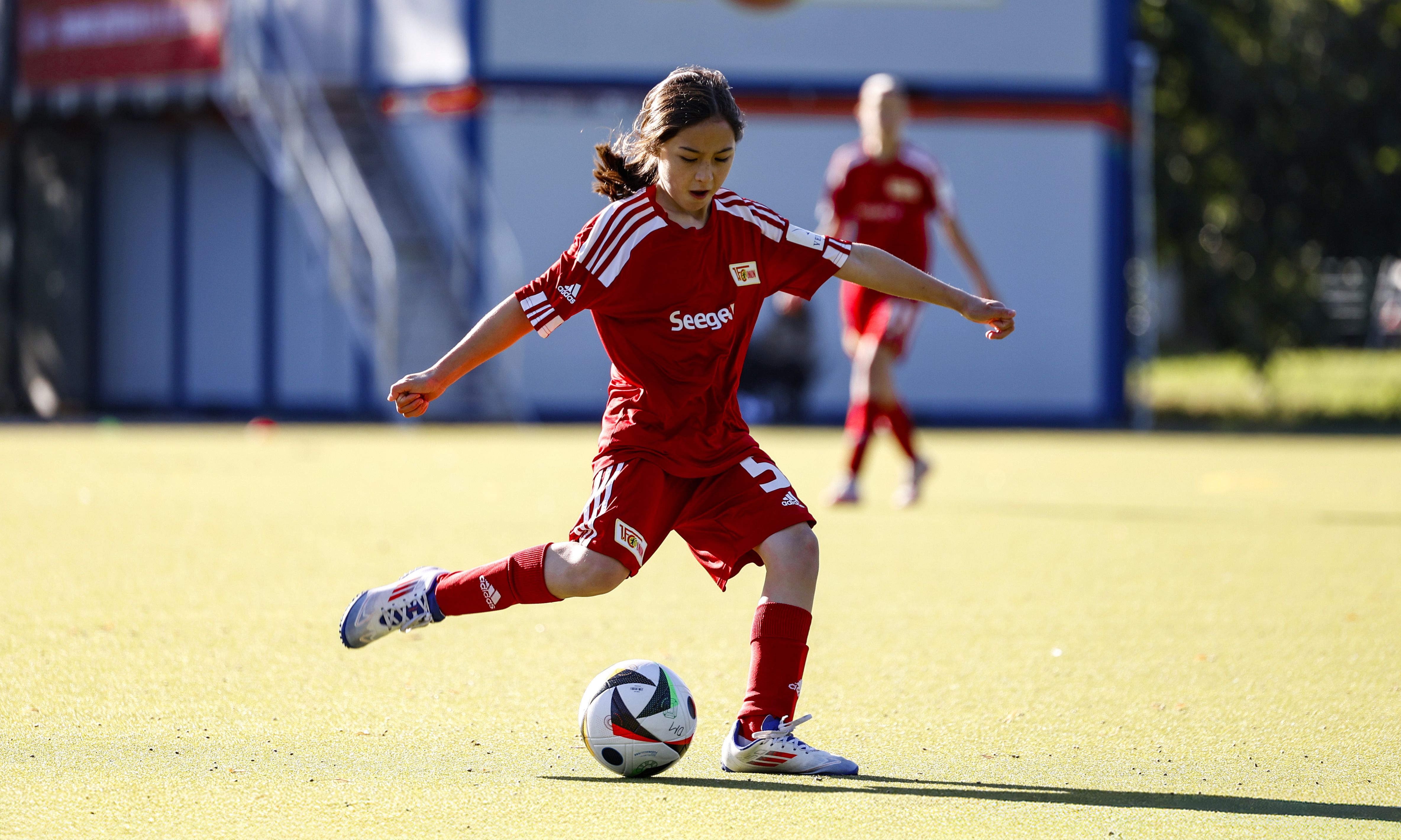 Ein Mädchen in rotem Fußballtrikot tritt einen Ball auf einem Tennisplatz. Im Hintergrund sind weitere Spielerinnen.