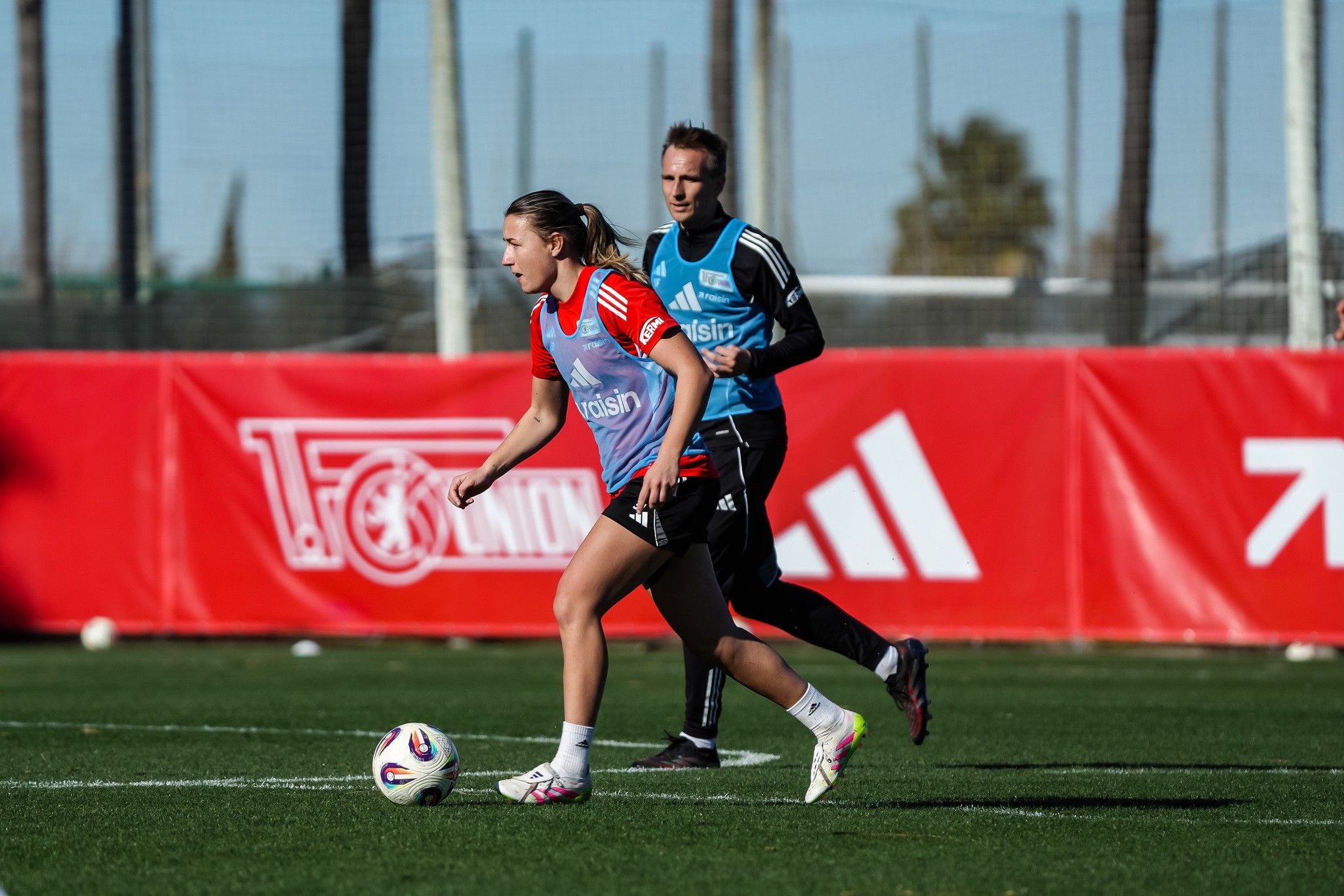 Eine Fußballspielerin übt auf einem Trainingsplatz, während ein Trainer hinter ihr steht. Rote und grüne Trainingstrikots sind zu sehen.
