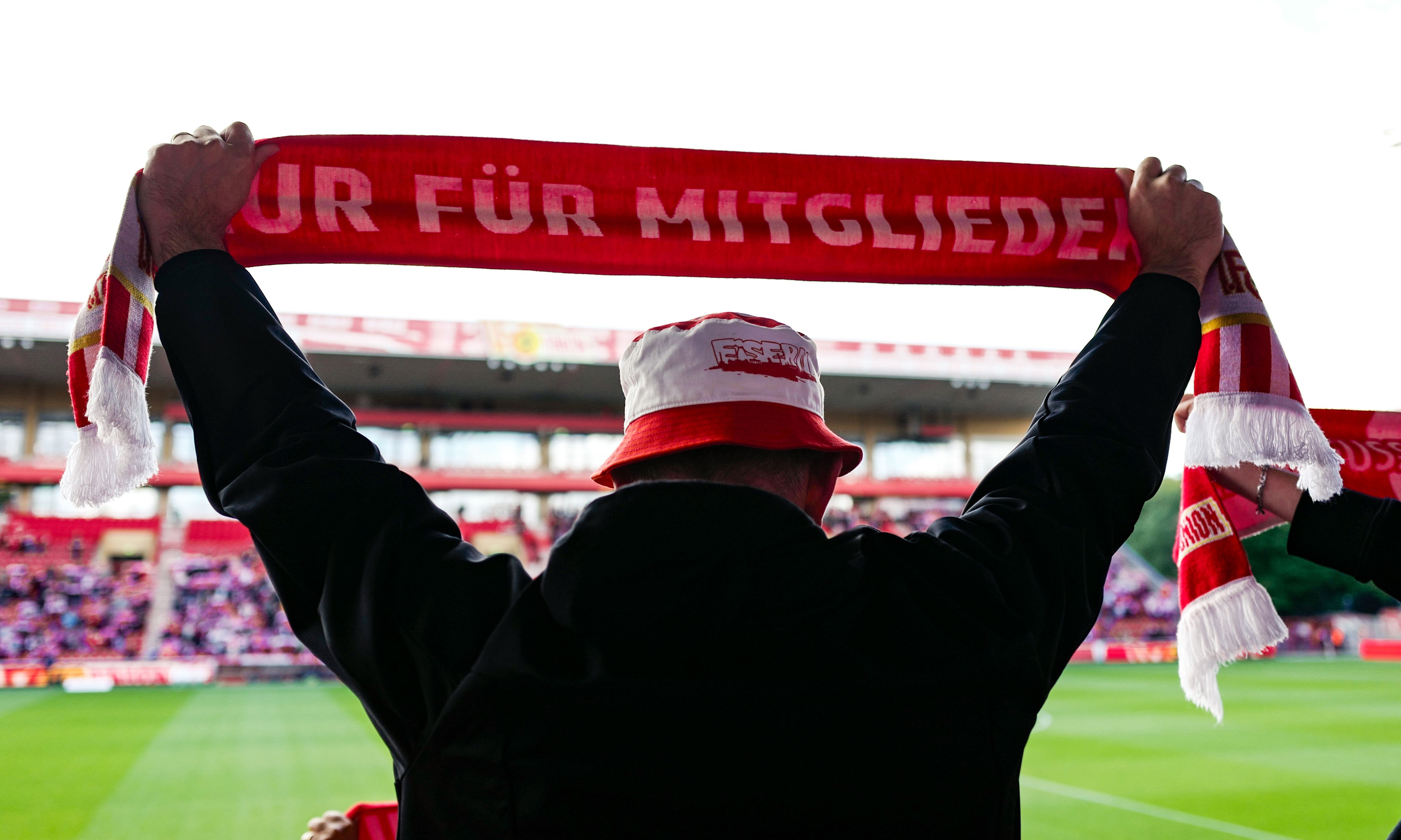 Ein Fan hält einen roten Schal mit weißem Schriftzug hoch, während im Hintergrund ein Fußballspiel stattfindet.