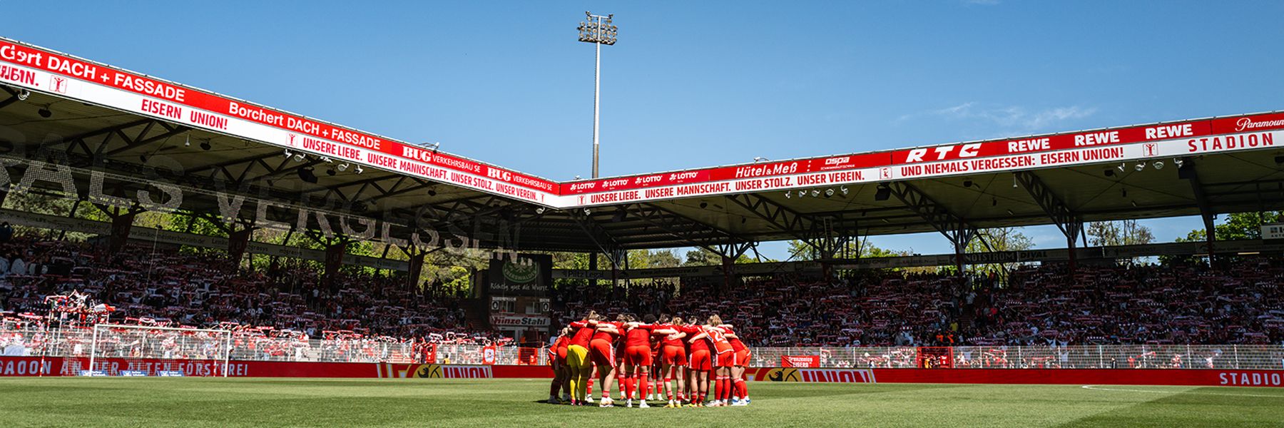 Players of a football team stand in a circle discussing in a stadium with spectators and advertising banners.