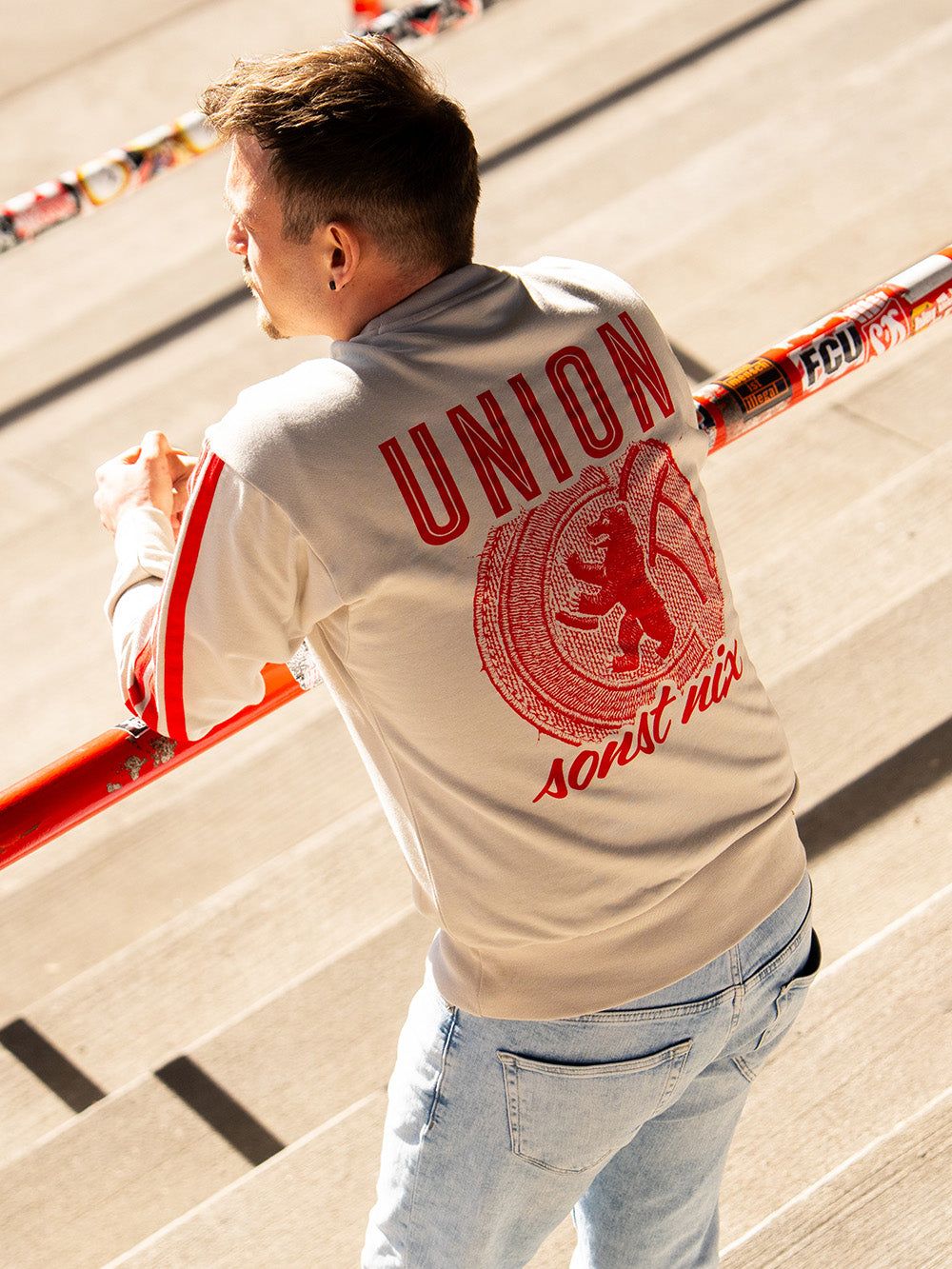 Mann in einem beige-roten Sweatshirt mit der Aufschrift "UNION" steht an einer Treppe mit bunten Handläufen.
