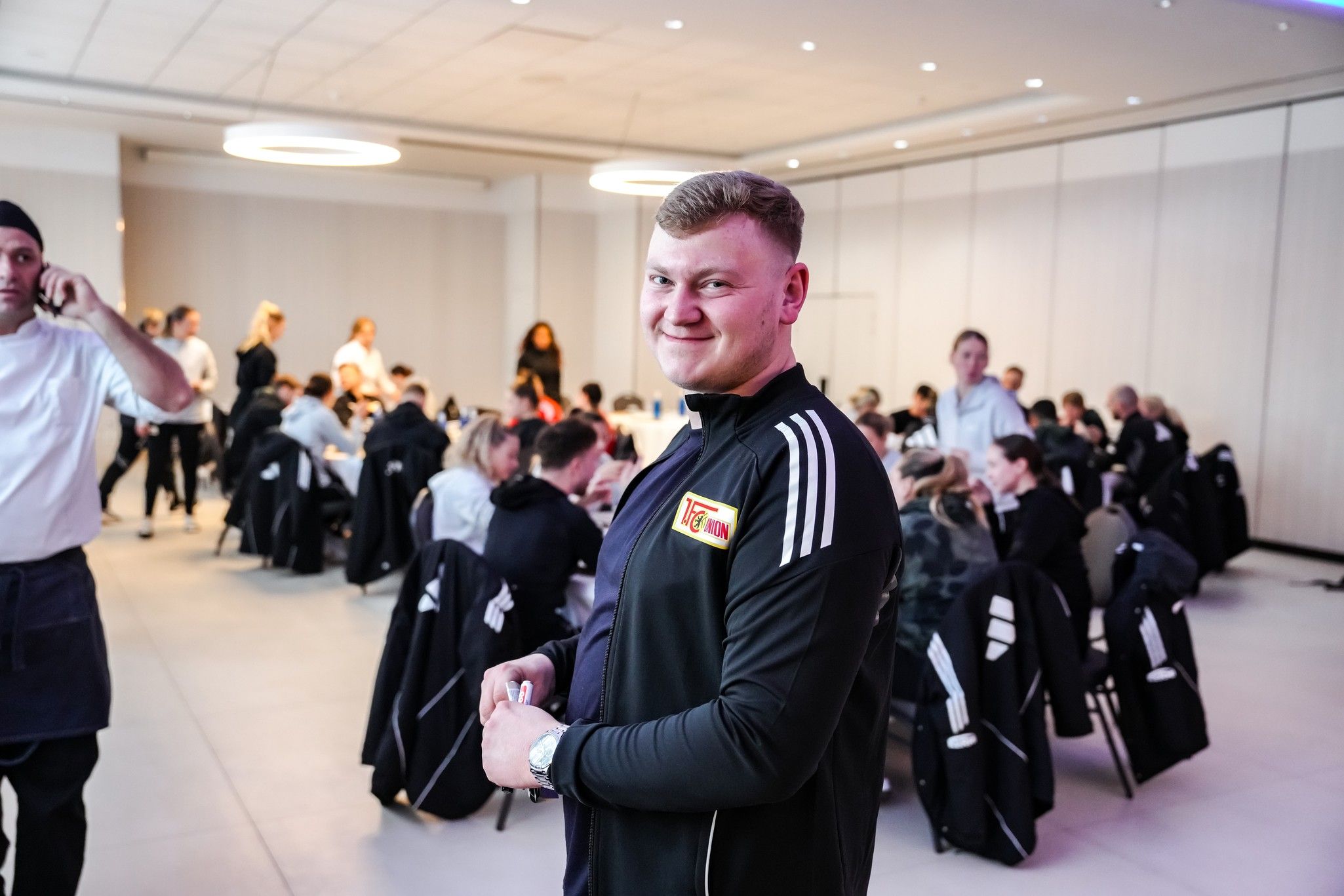 Ein Mann in einem schwarzen Sportjackett steht lächelnd in einem Raum mit vielen anderen Personen, die an Tischen sitzen.