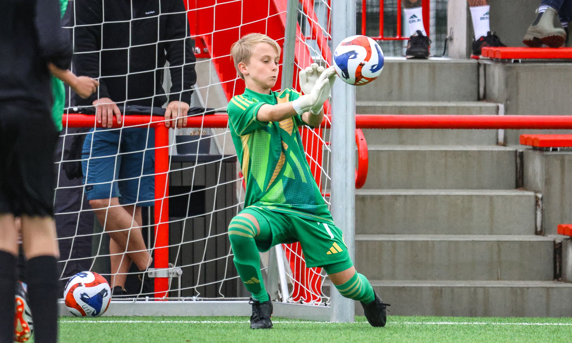 Ein junger Torwart in grüner Sportkleidung fängt einen Fußball während eines Spiels. Im Hintergrund sind Zuschauer und eine Torlinie sichtbar.