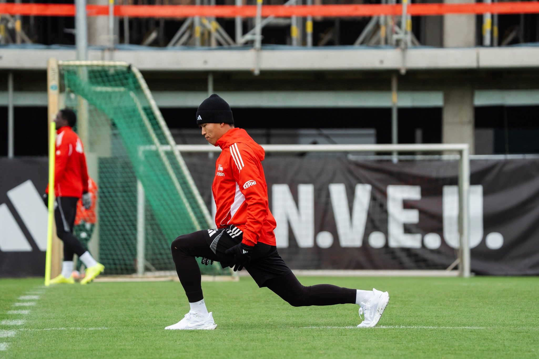 Fußballspieler in rot-schwarzer Trainingskleidung, der sich auf einem Platz dehnt, im Hintergrund eine Baustelle.