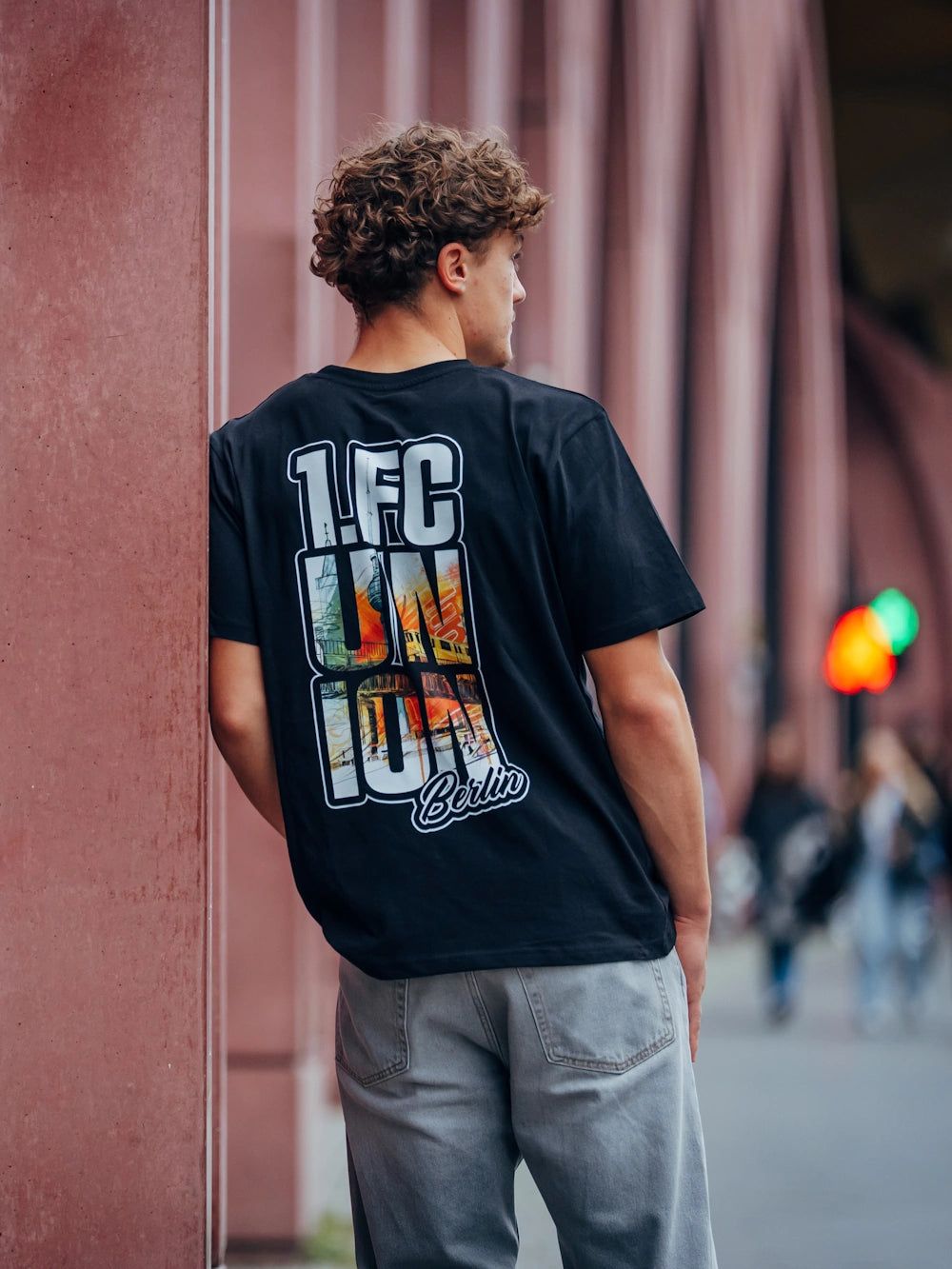 Junger Mann trägt ein schwarzes T-Shirt mit dem Aufdruck "1. FC Union Berlin" und steht vor einem urbanen Hintergrund.