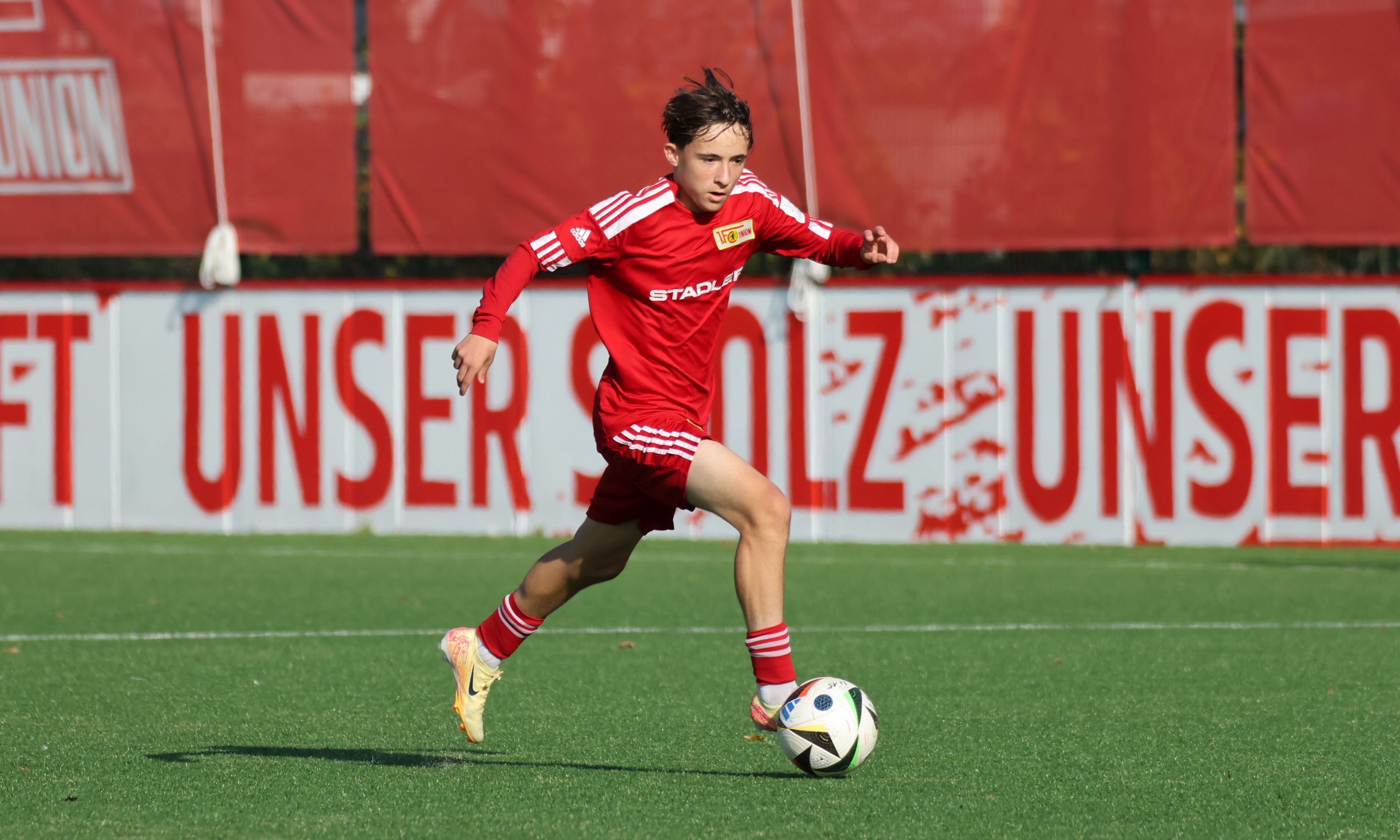 Ein junger Fußballspieler in rotem Trikot sprintet auf einem Sportfeld mit einem Ball. Im Hintergrund steht ein Banner mit dem Text „UNSER SOlz“.