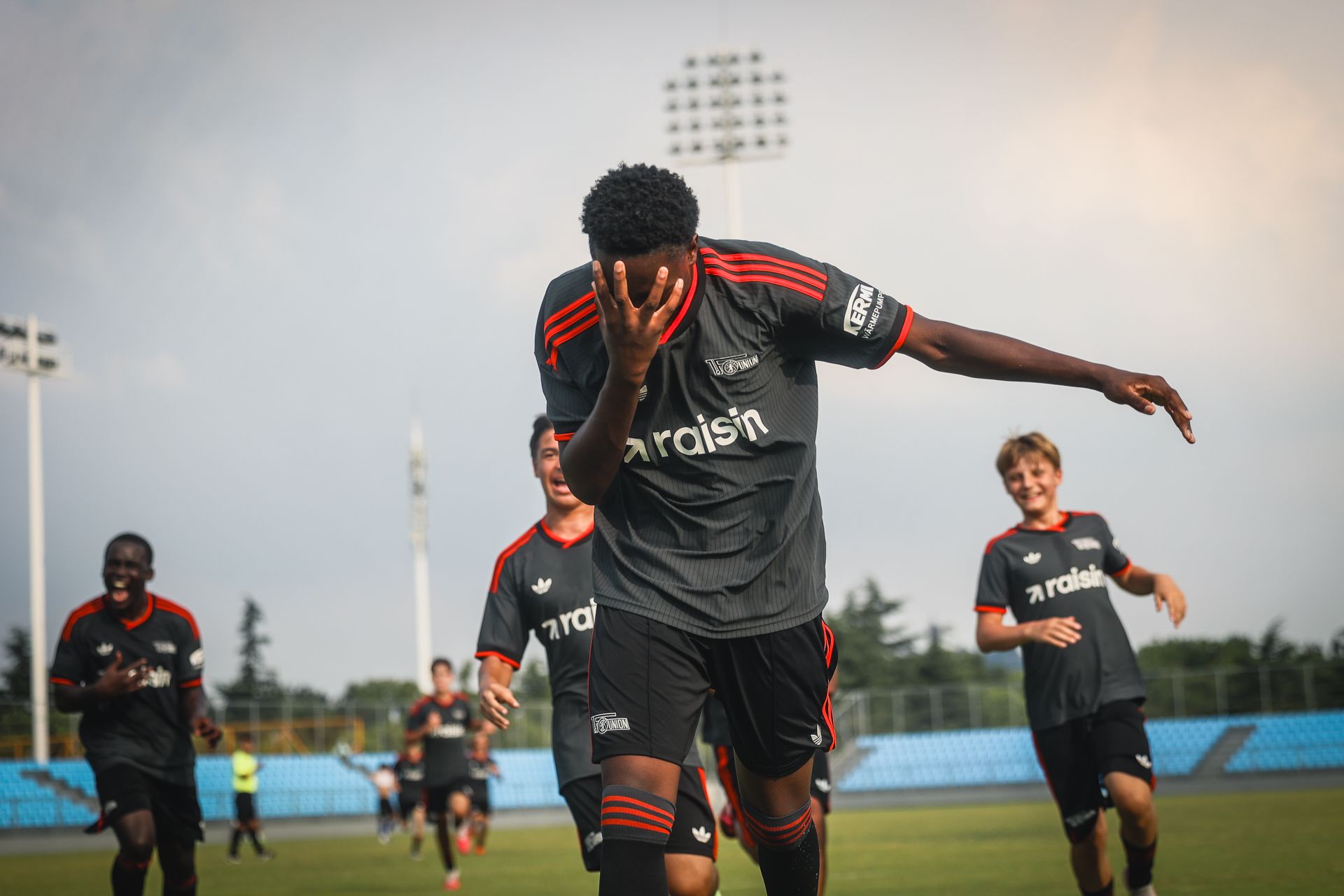 Ein Fußballspieler jubelt nach einem Tor, während Teamkollegen im Hintergrund feiern. Es ist ein sonniger Tag auf dem Trainingsplatz.