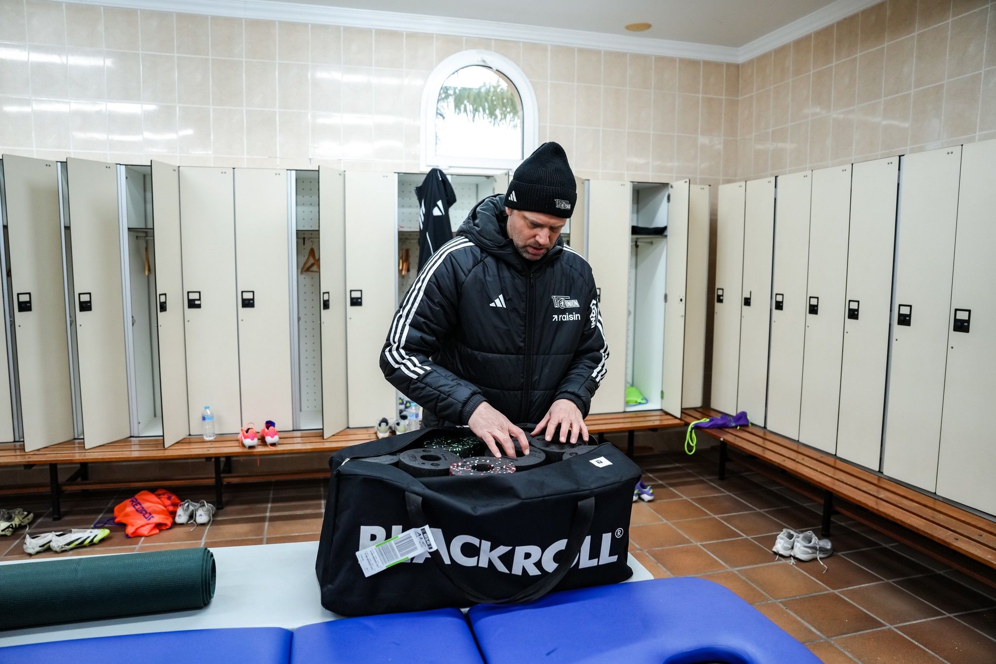 Ein Sportler steht in einem Umkleideraum und packt eine große Tasche mit Ausrüstung aus. Numerierte Schränke im Hintergrund.