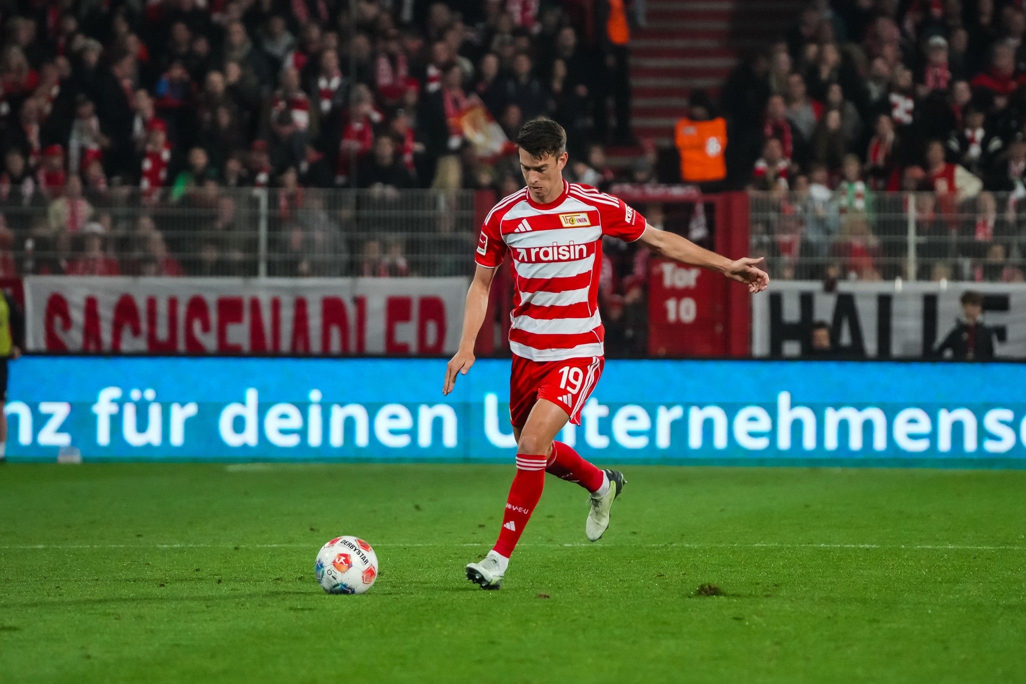 Ein Fußballspieler in einem rot-weiß gestreiften Trikot führt einen Freistoß aus, während Zuschauer im Hintergrund jubeln.
