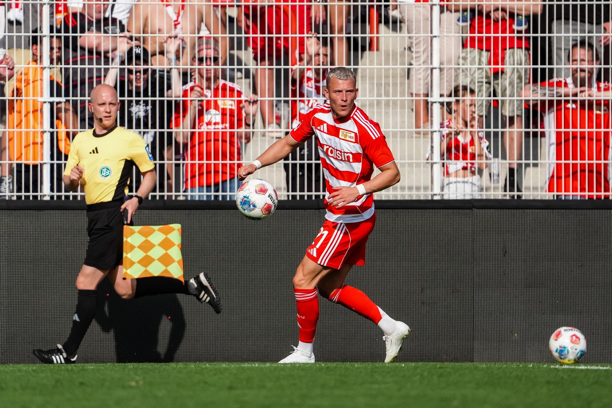 Ein Fußballspieler in rotem Trikot mit weißen Details steht am Spielfeldrand, während ein Schiedsrichter ihn beobachtet.