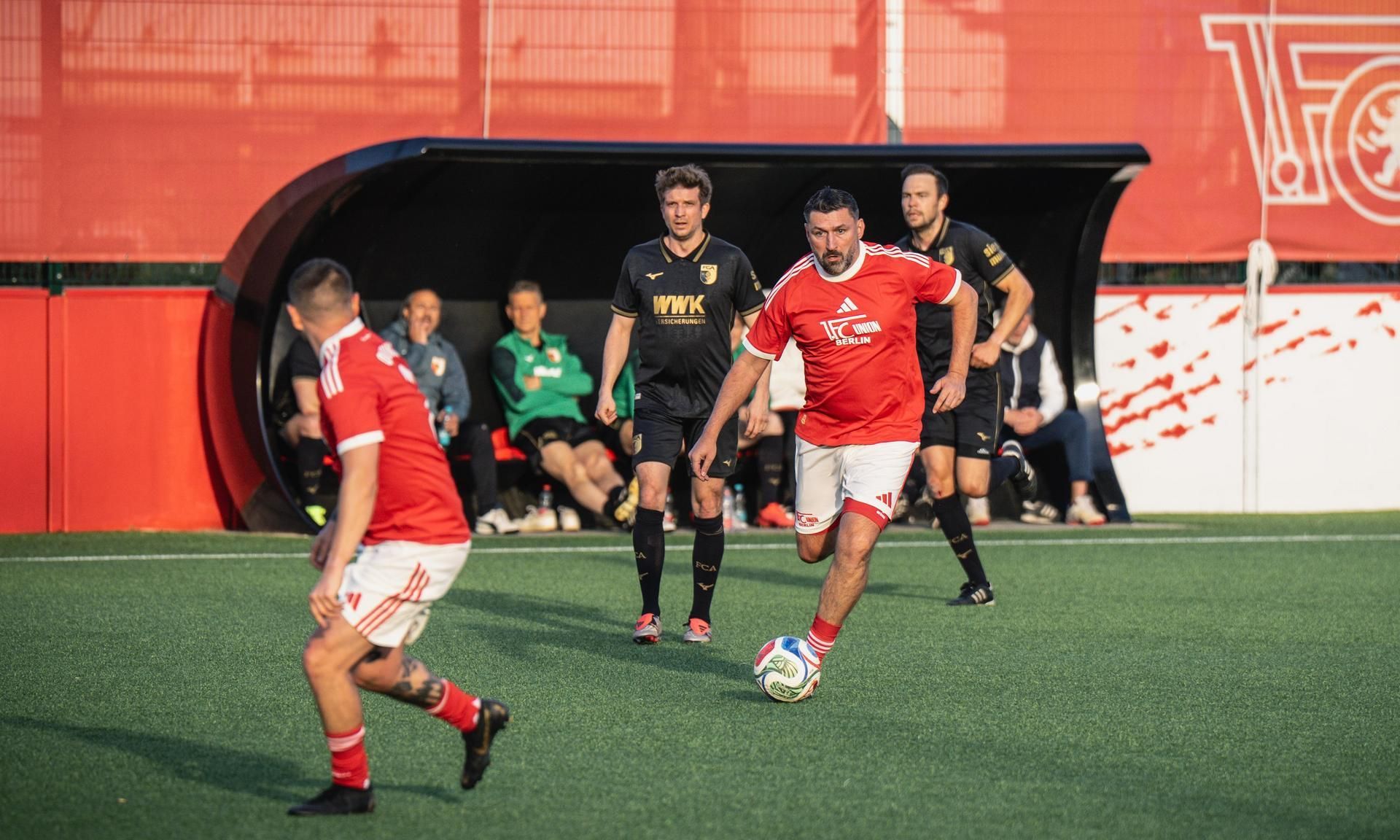 Ein Fußballspieler in rotem Trikot dribbelt mit dem Ball, während andere Spieler und Zuschauer im Hintergrund stehen.