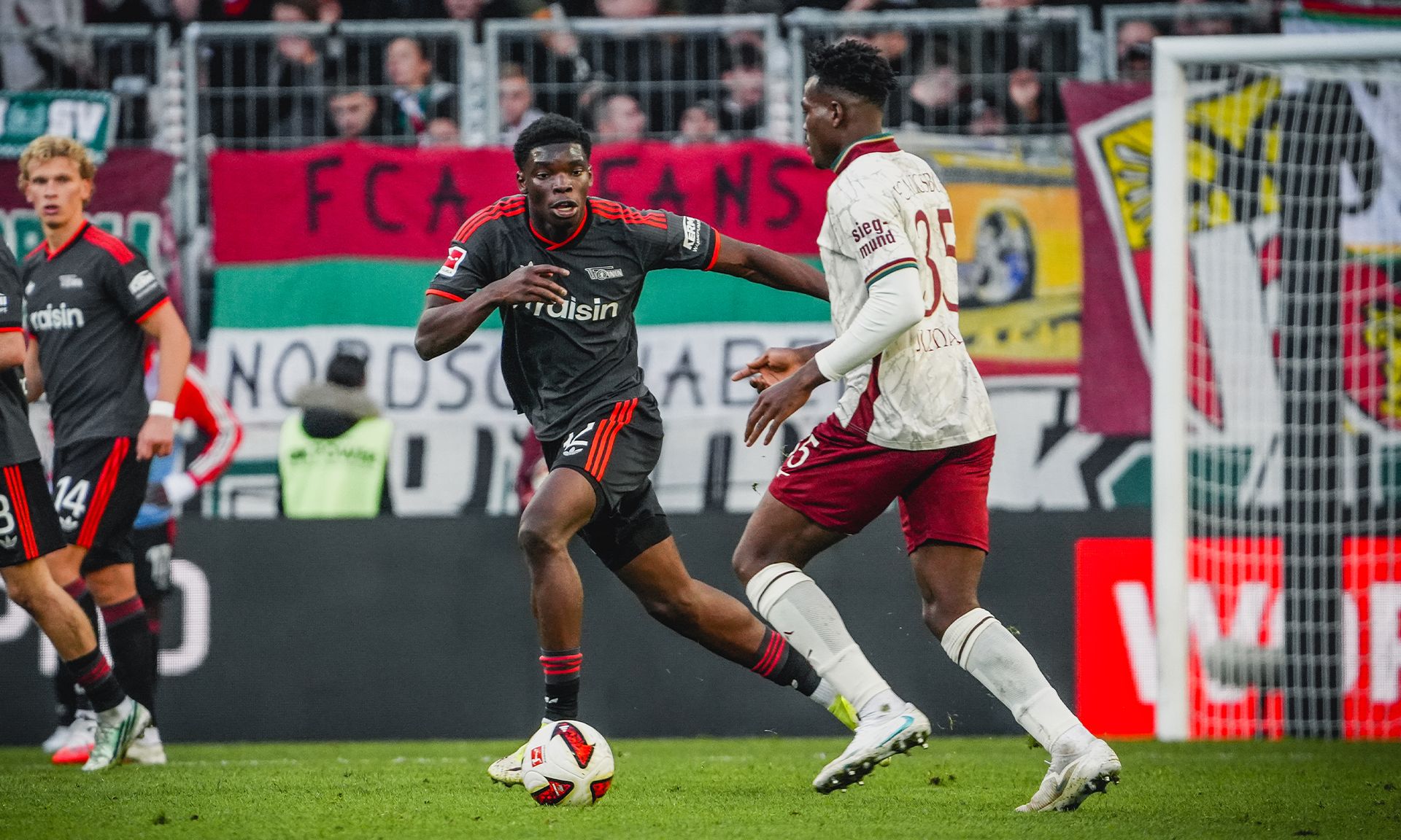 Zwei Fußballspieler stehen sich auf dem Spielfeld gegenüber, während im Hintergrund Fans und Banner zu sehen sind.