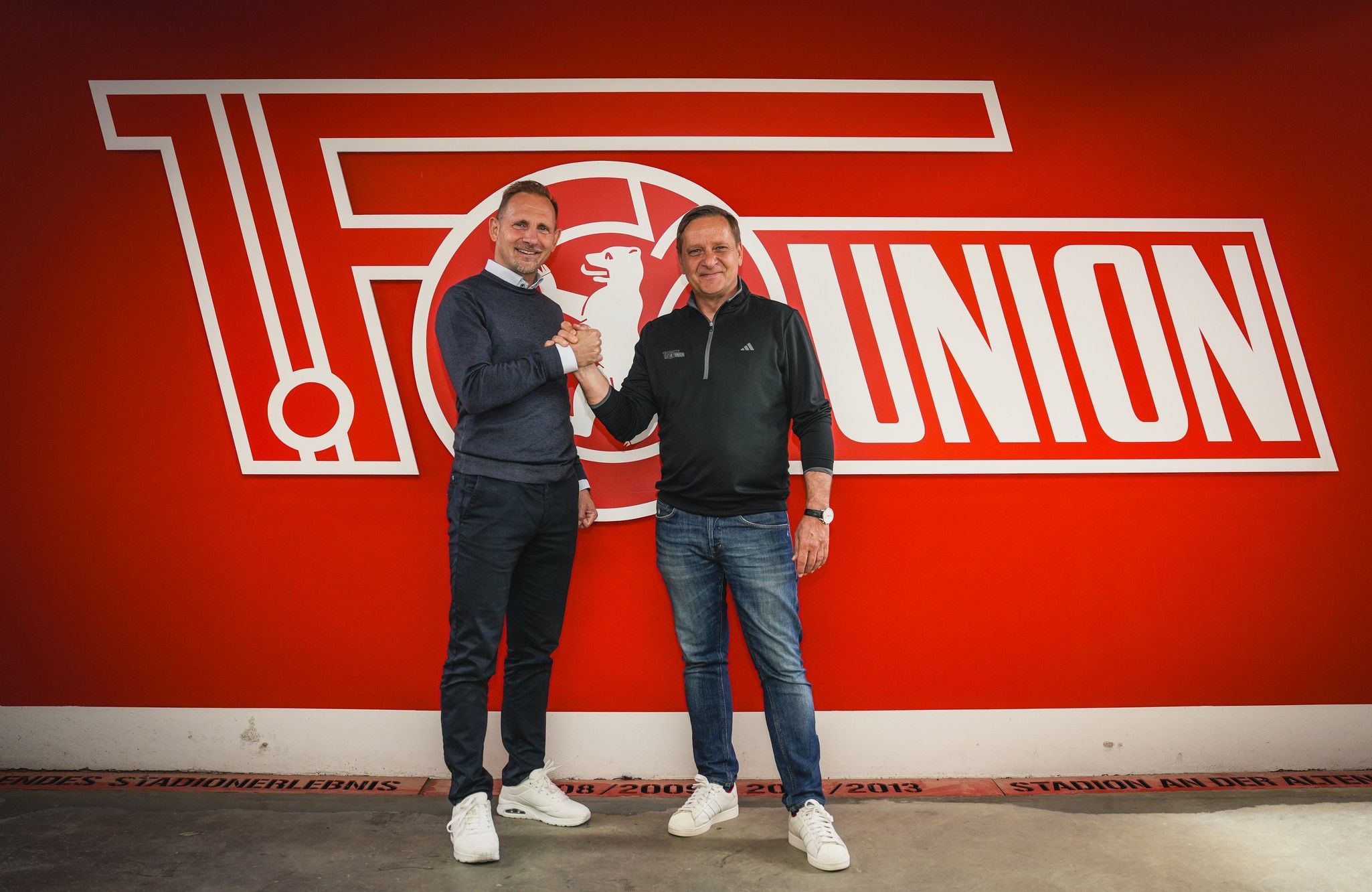 Zwei Männer schütteln sich die Hände vor einer roten Wand mit dem Logo von Union Berlin im Hintergrund.