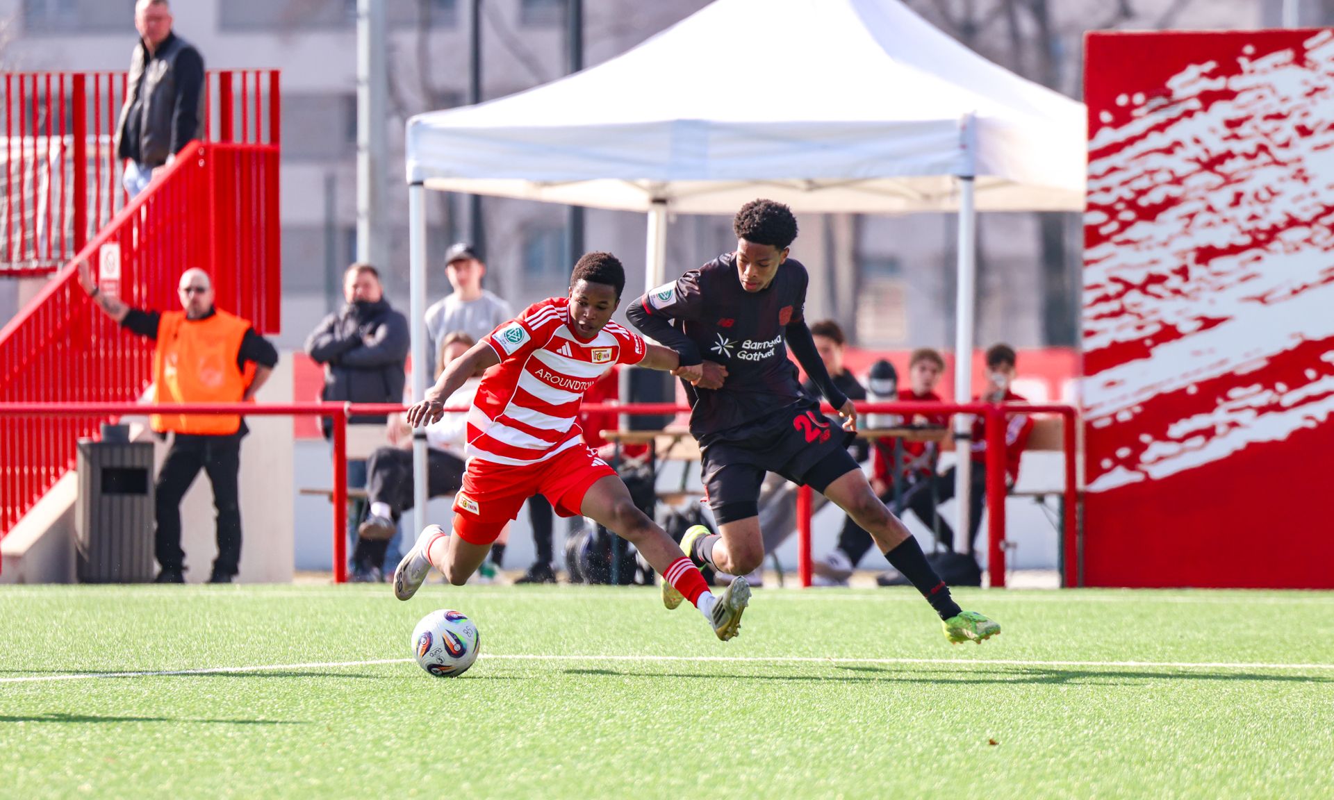 Zwei Fußballspieler im Wettkampf um den Ball auf einem Kunstrasenplatz, Zuschauer im Hintergrund.