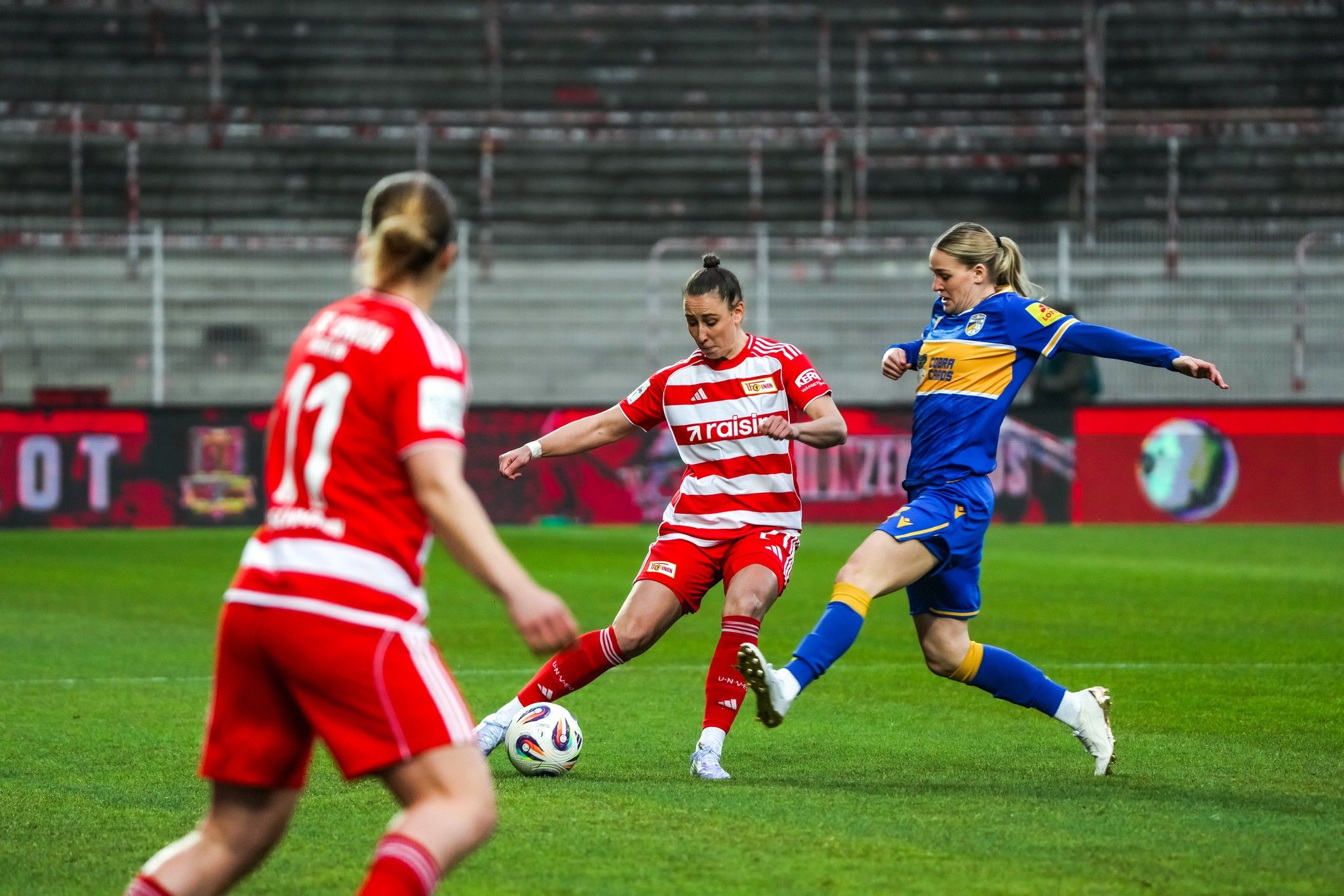 Drei Fußballspielerinnen im Wettkampf auf dem Spielfeld, eine in roten Trikots und zwei in blauen Trikots.