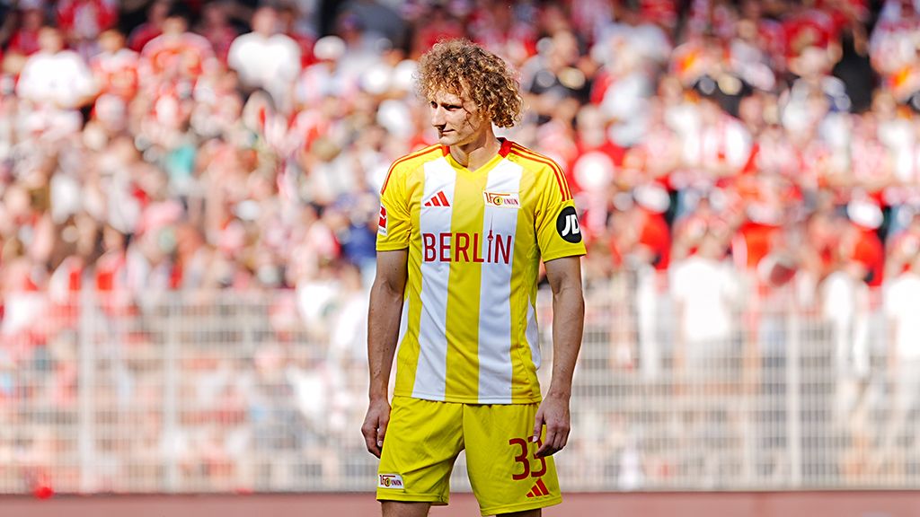 A soccer player in yellow and white uniform stands on the field, surrounded by a cheering crowd.