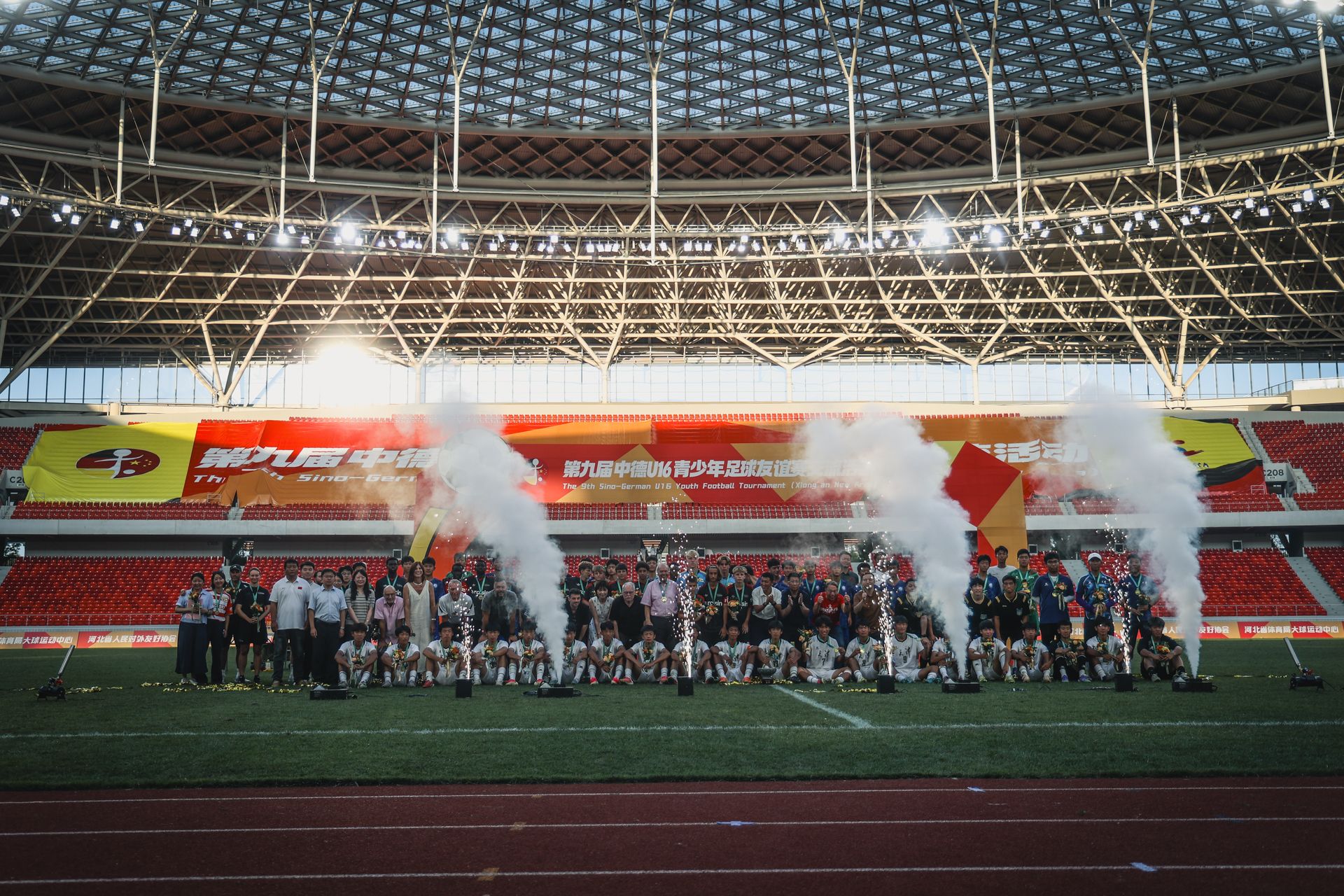 Eine große Gruppe von Menschen posiert auf einem Sportfeld, umgeben von bunten Bannern und Rauchern in einem Stadionsetting.