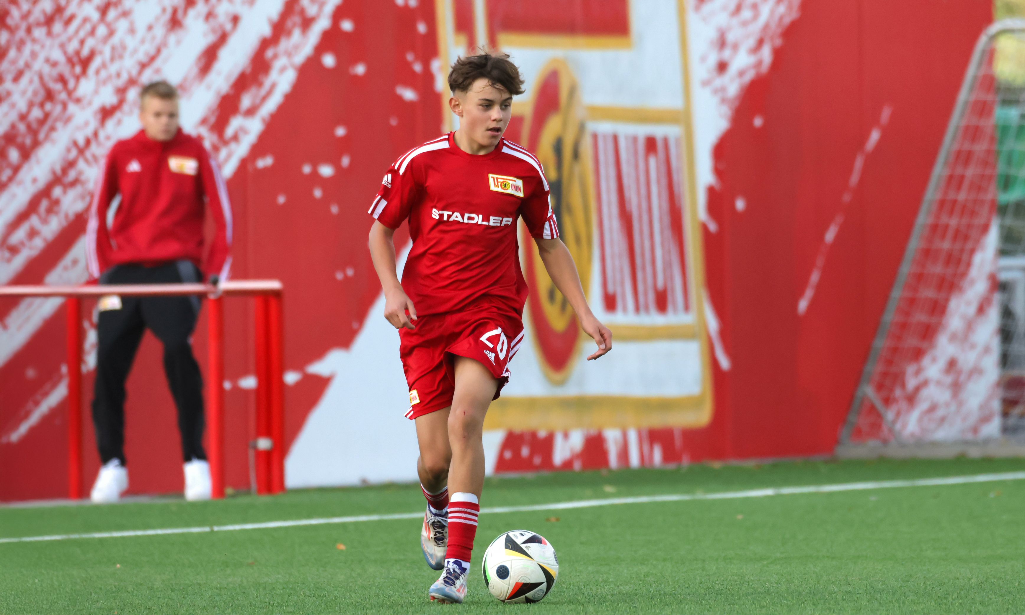 Ein junger Fußballspieler in rotem Trikot dribbelt den Ball auf einem Trainingsplatz. Im Hintergrund sind Vereinsfarben und ein weiterer Spieler sichtbar.