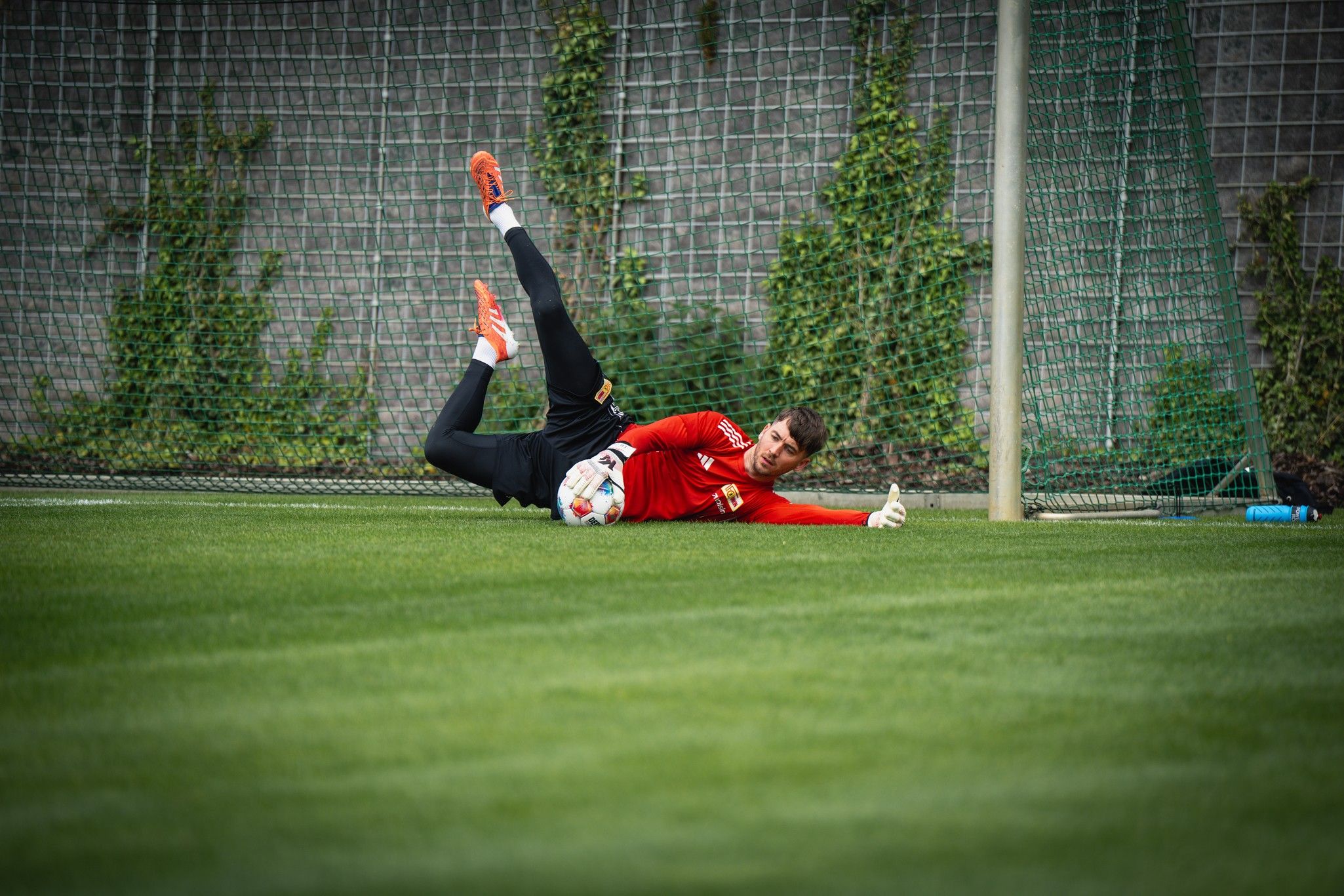 Ein Fußballtorwart in rotem Trikot stoppt den Ball während des Trainings auf einem Rasenfeld.