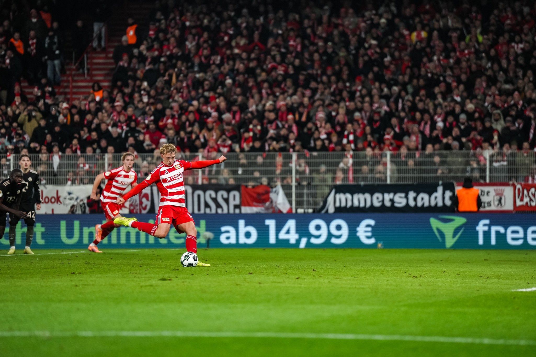 Fußballspieler in rot-weißer Uniform tritt den Ball auf einem Stadionrasen, Zuschauer im Hintergrund, Emotionen im Spiel.
