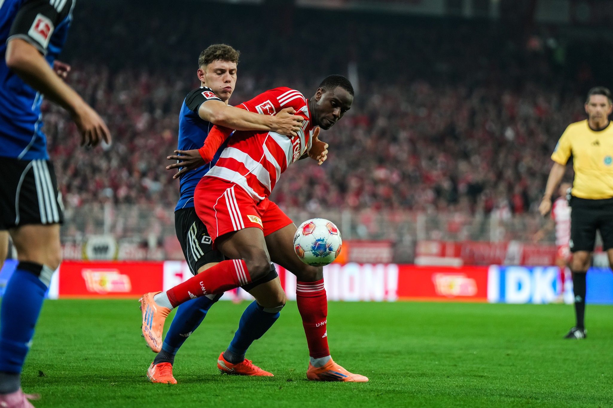Zwei Fußballspieler kämpfen um den Ball auf einem Stadionfeld, Zuschauer im Hintergrund.