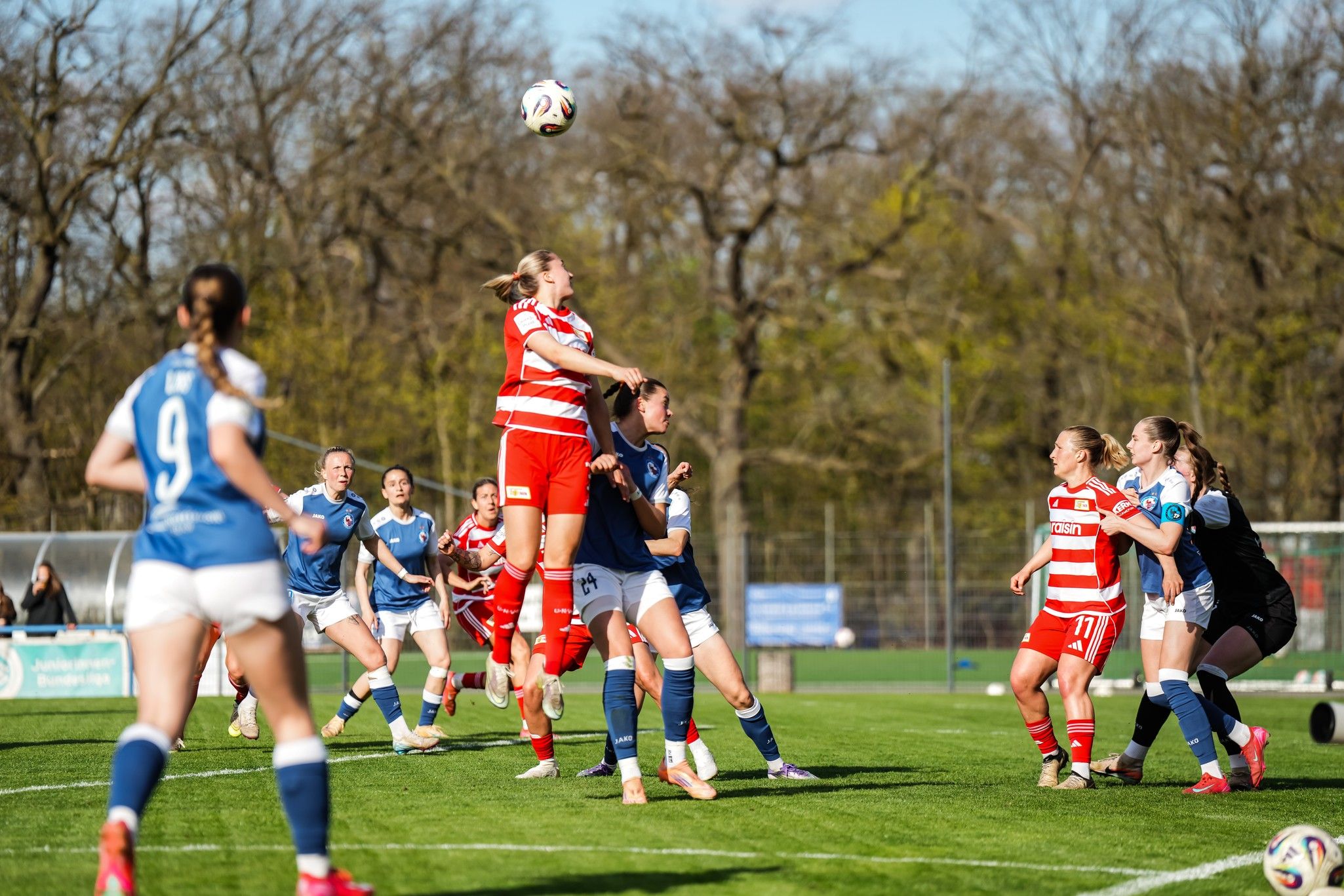 Eine Fußballspielszene mit weiblichen Spielern in Aktion, während eine Spielerin hochspringt, um den Ball zu erreichen.