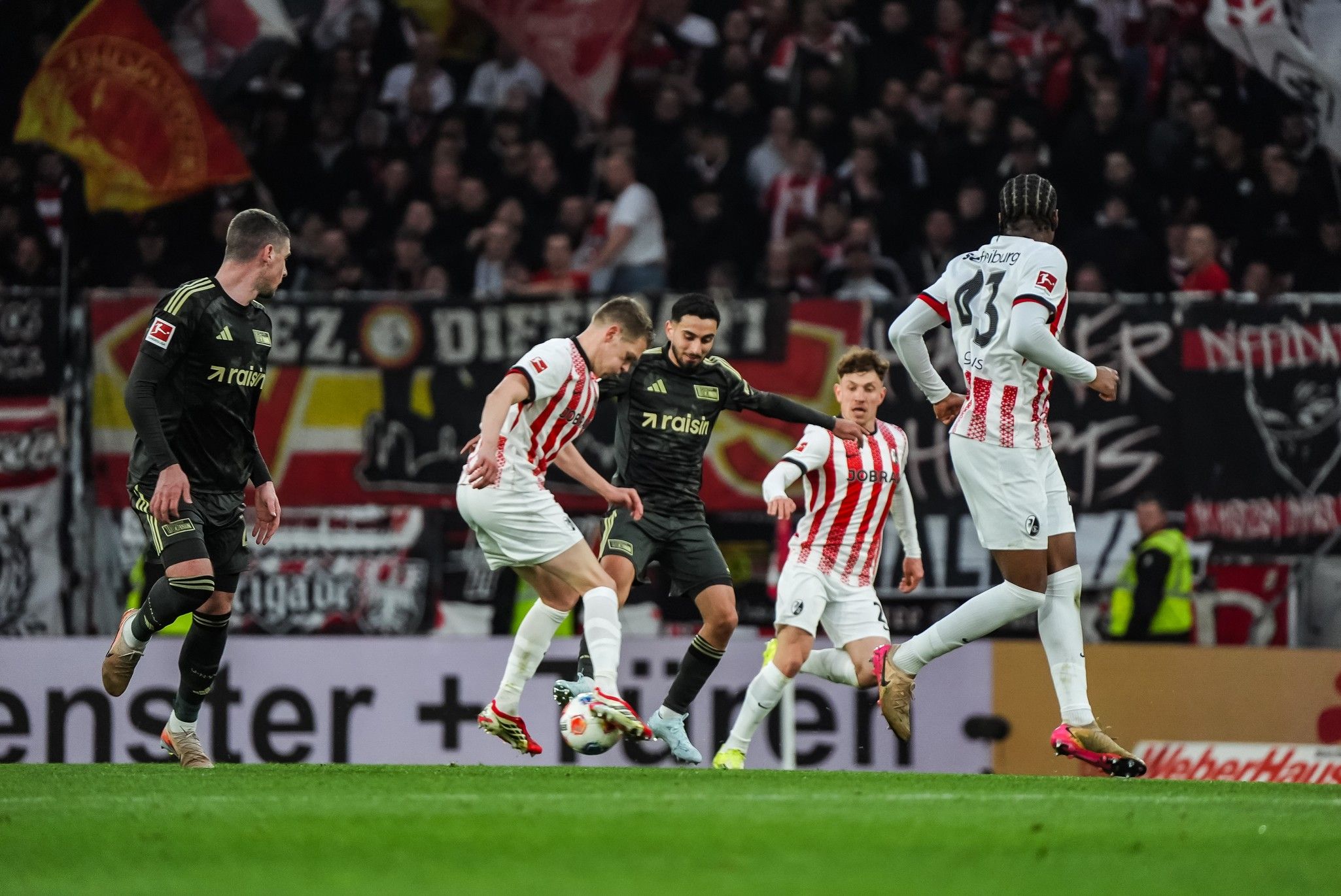Ein Fußballspiel mit Spielern in roten und weißen Trikots, die auf einem Stadionplatz agieren, Umrahmung von Fan-Transparenten.