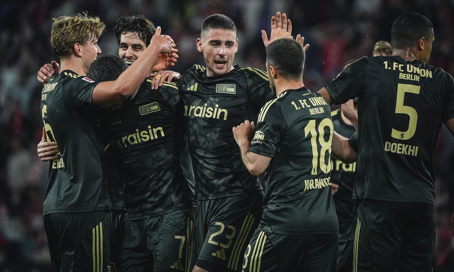 Fußballspieler des 1. FC Union Berlin feiern ein Tor, umgeben von jubelnden Teamkollegen in schwarzen Trikots.