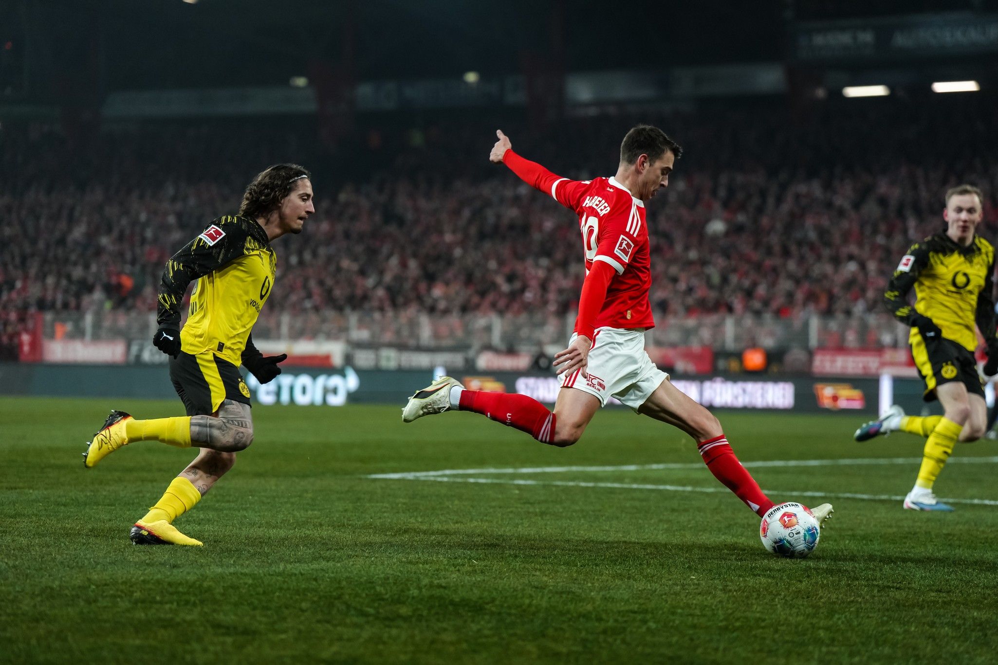 Ein Fußballspieler in rotem Trikot dribbelt den Ball, während ein Spieler in schwarzem Trikot ihm folgt. Im Hintergrund eine große Menschenmenge.