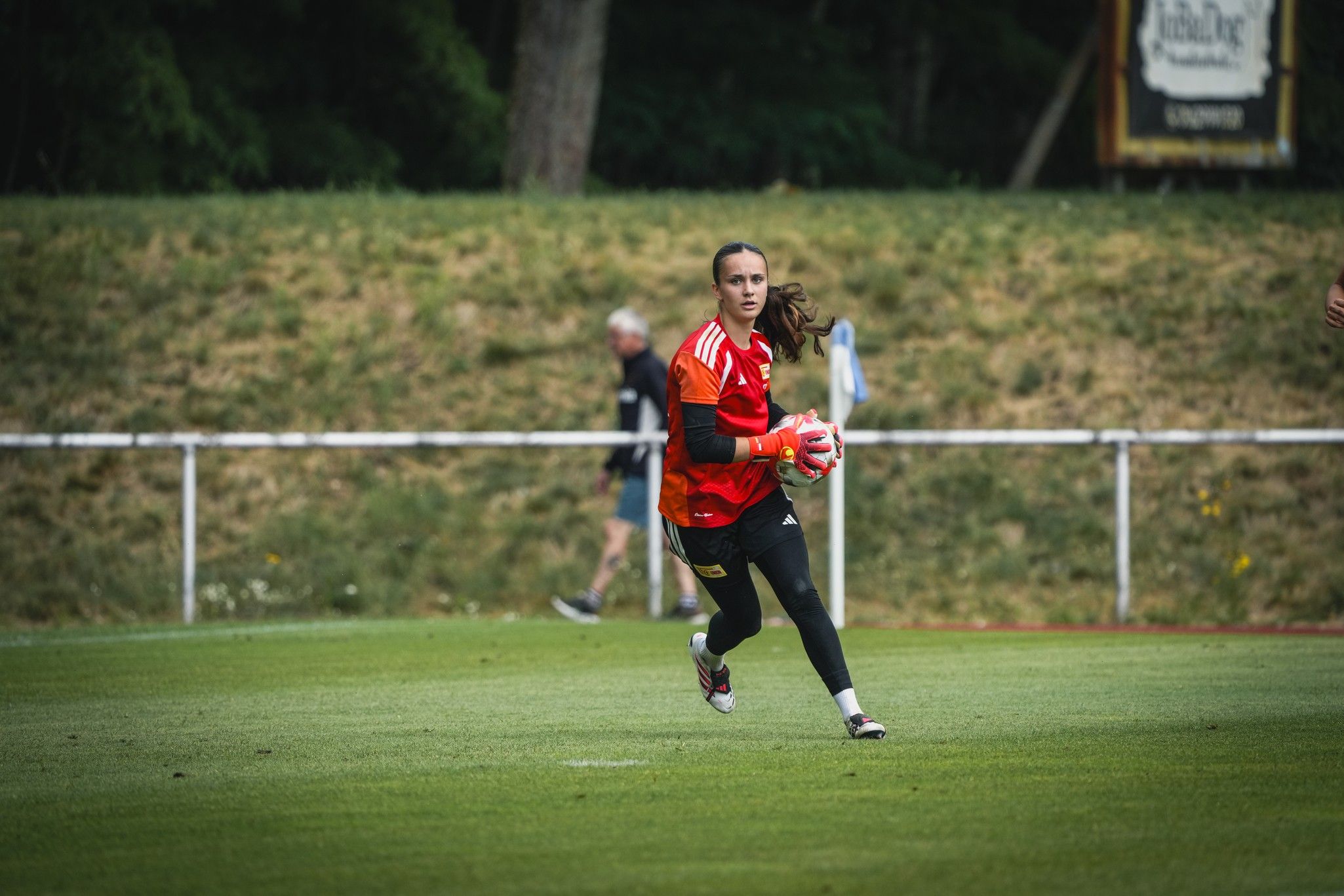 Eine junge Frau in einem roten Trikot und schwarzen Hosen läuft mit einem Fußball auf einem grünen Sportplatz.