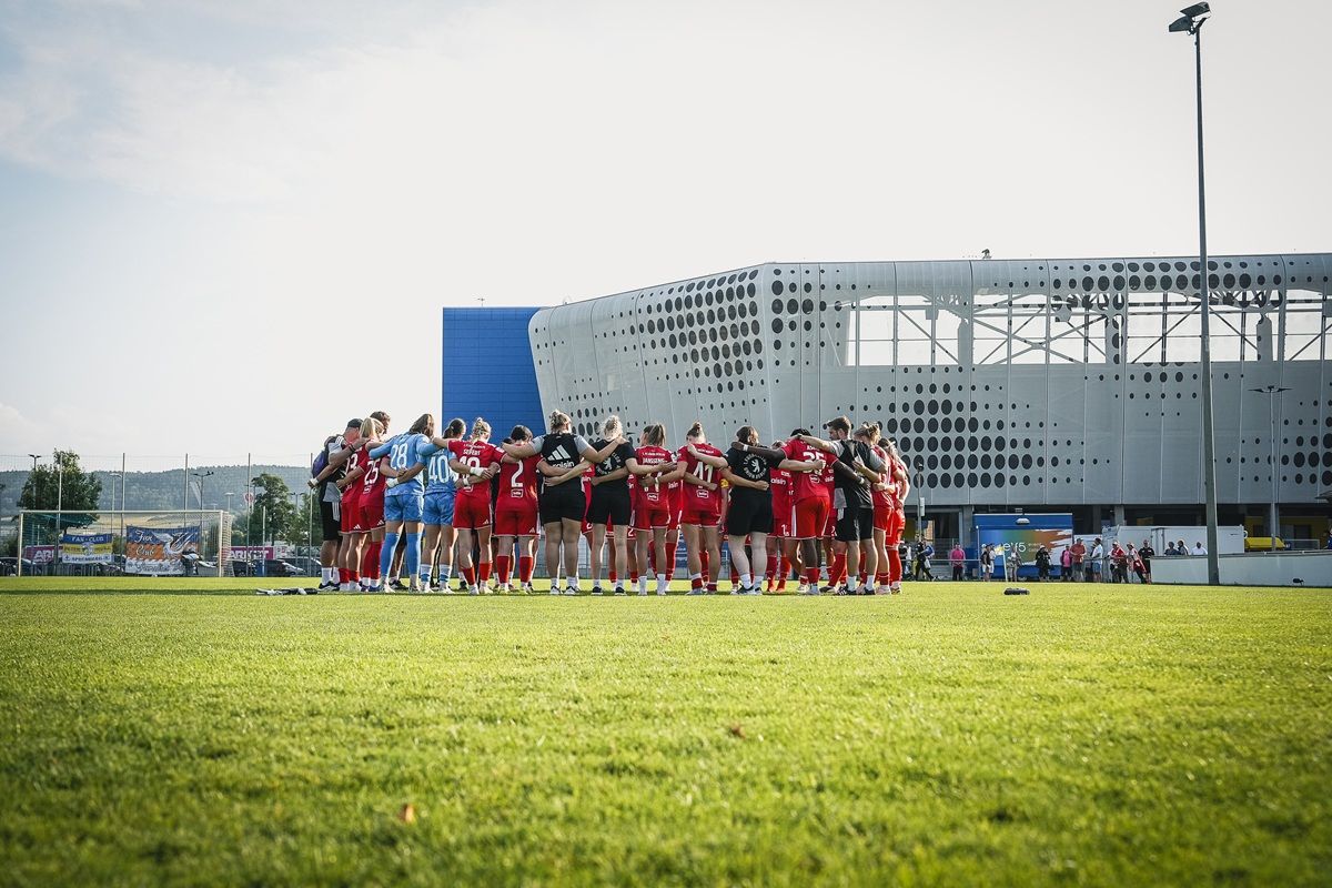 Gruppierung von Sportlern in roten Trikots, die sich auf einem Fußballfeld umarmen, vor einem modernen Gebäude im Hintergrund.