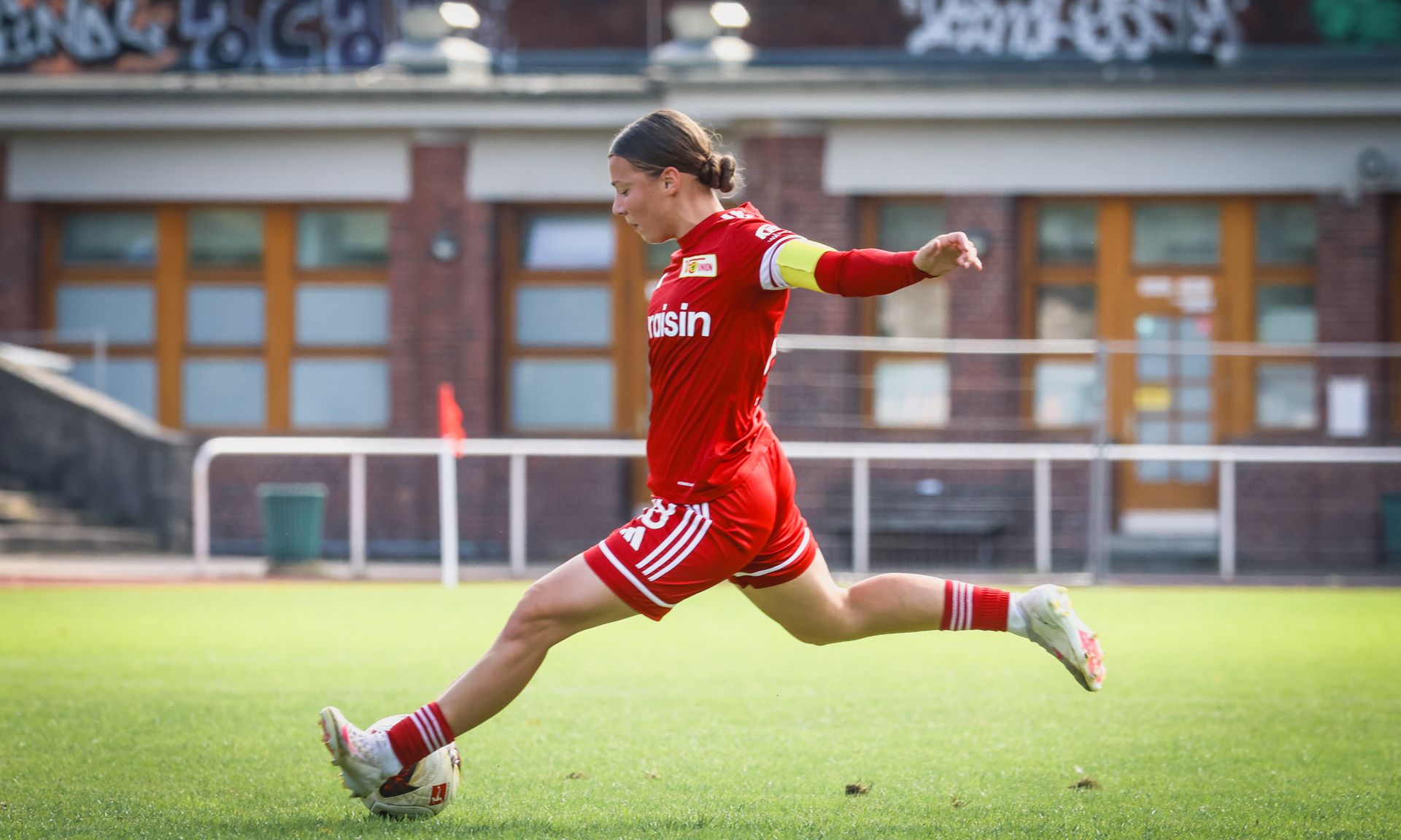 Eine Frauenfußballspielerin in roter Uniform tritt den Ball auf einem grünen Sportplatz.