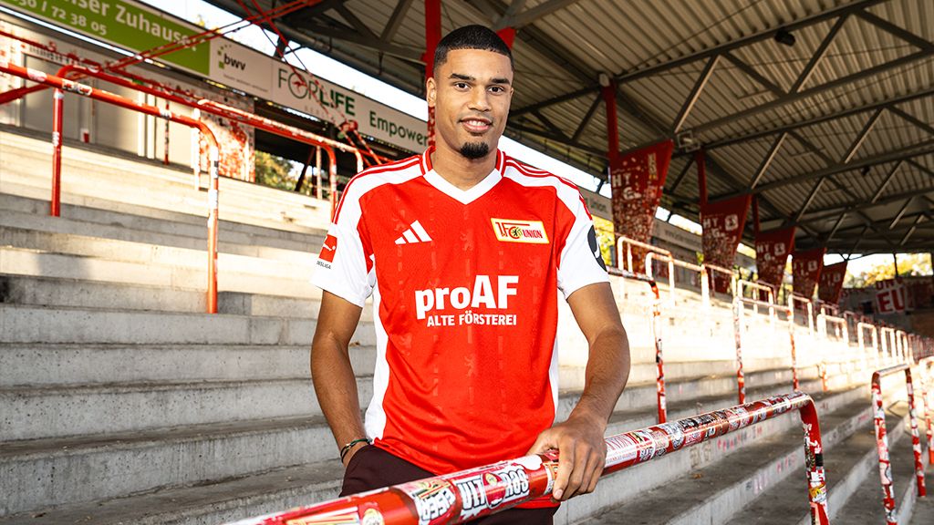 Ein junger Fußballspieler in einem rot-weißen Trikot steht an einer Tribüne im Stadion und lächelt.