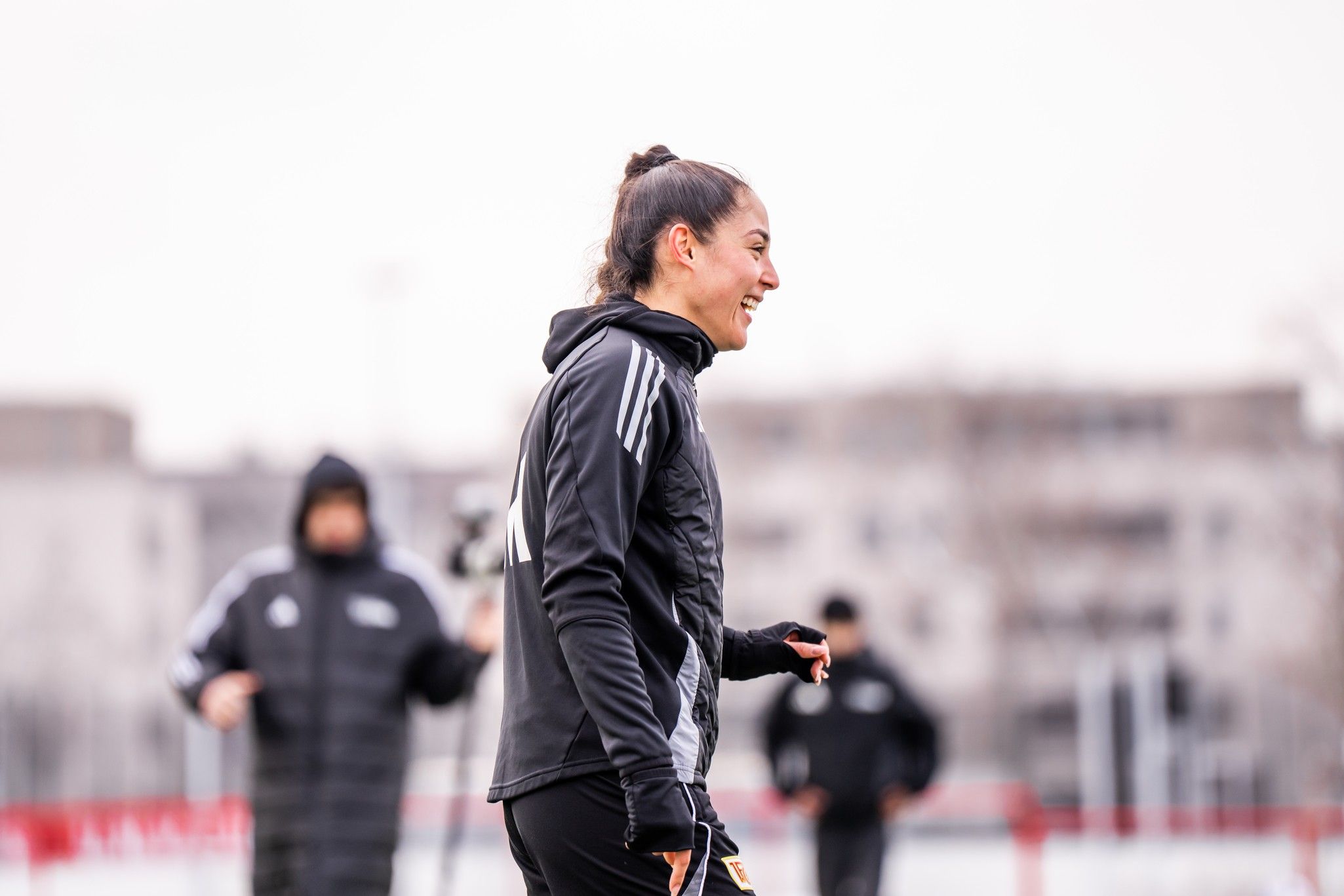 Eine Fußballspielerin lächelt während des Trainings auf dem Platz, im Hintergrund sind weitere Personen zu sehen.