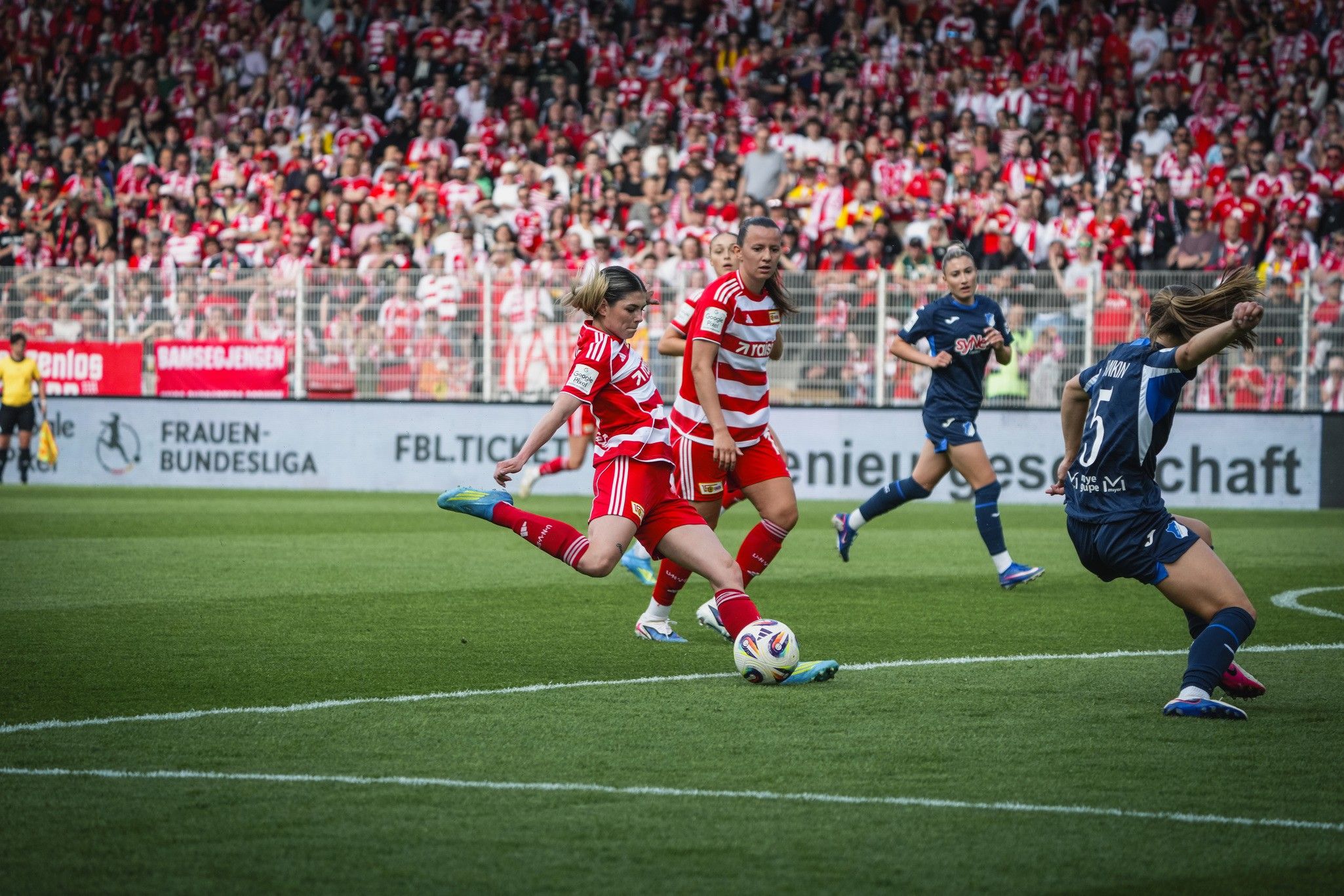 Eine Fußballspielerin in rotem Trikot schießt den Ball vor einer jubelnden Menge im Hintergrund.
