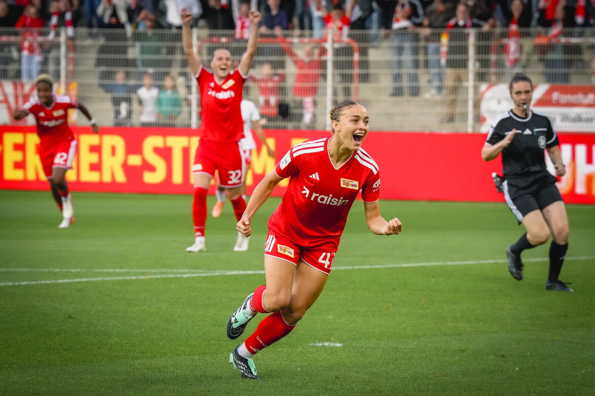 Eine Spielerin in rotem Trikot feiert nach einem Tor, während Teamkollegen im Hintergrund jubeln. Schiedsrichterin beobachtet die Szene.