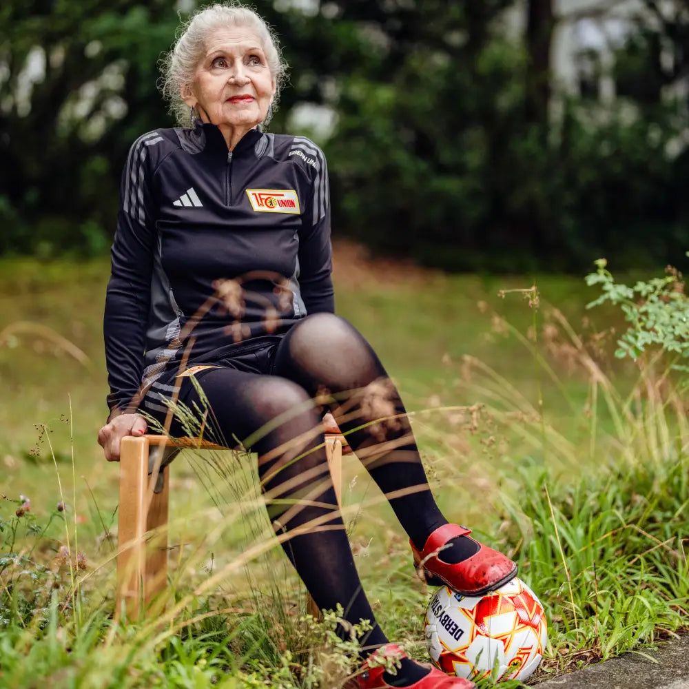 Ältere Frau mit grauen Haaren sitzt auf einem Stuhl im Freien, trägt Sportkleidung und hat einen Fußball neben sich.