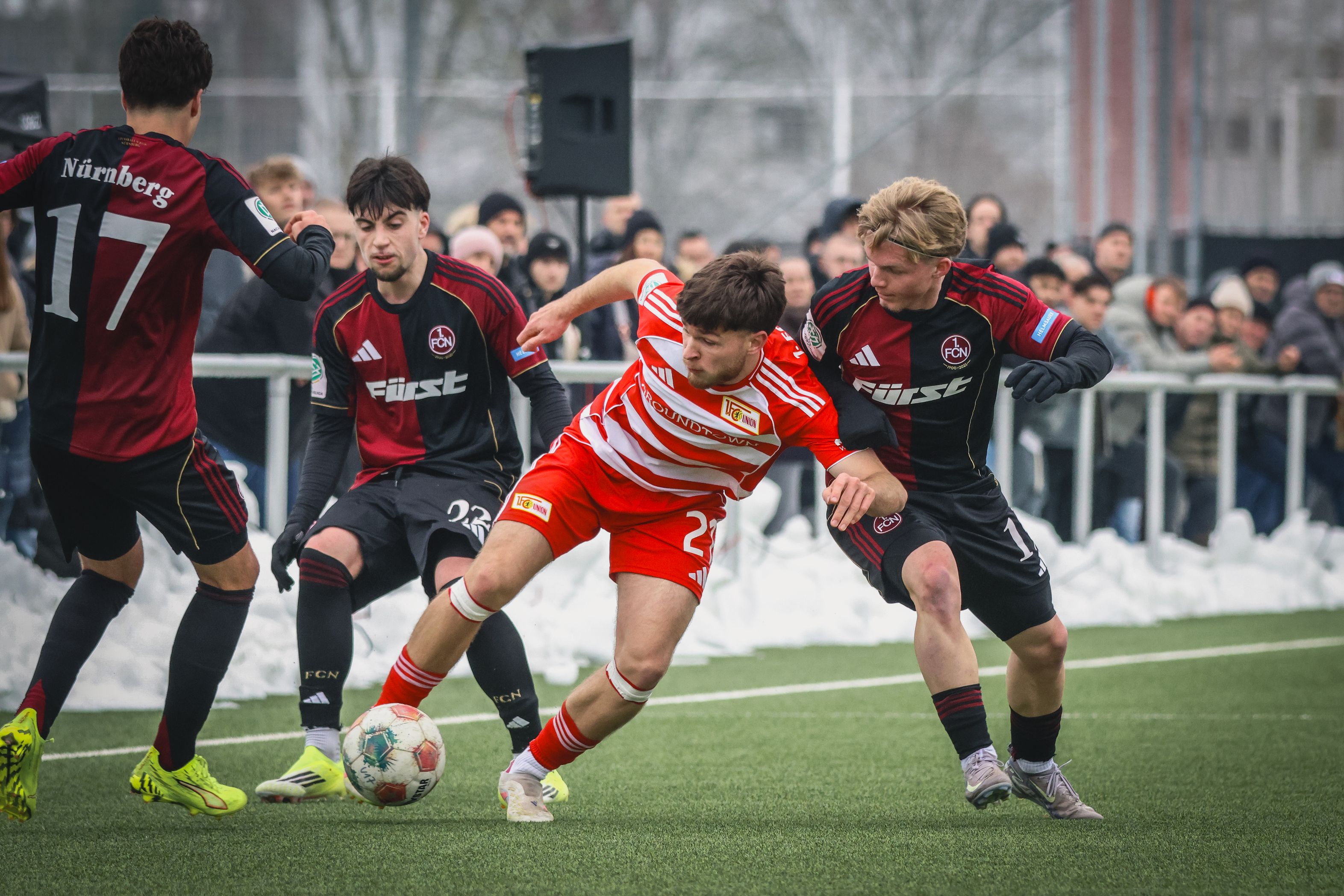 Ein Spieler in gestreiftem Trikot dribbelt während eines Fußballspiels, umgeben von zwei Verteidigern auf einem grünen Feld.