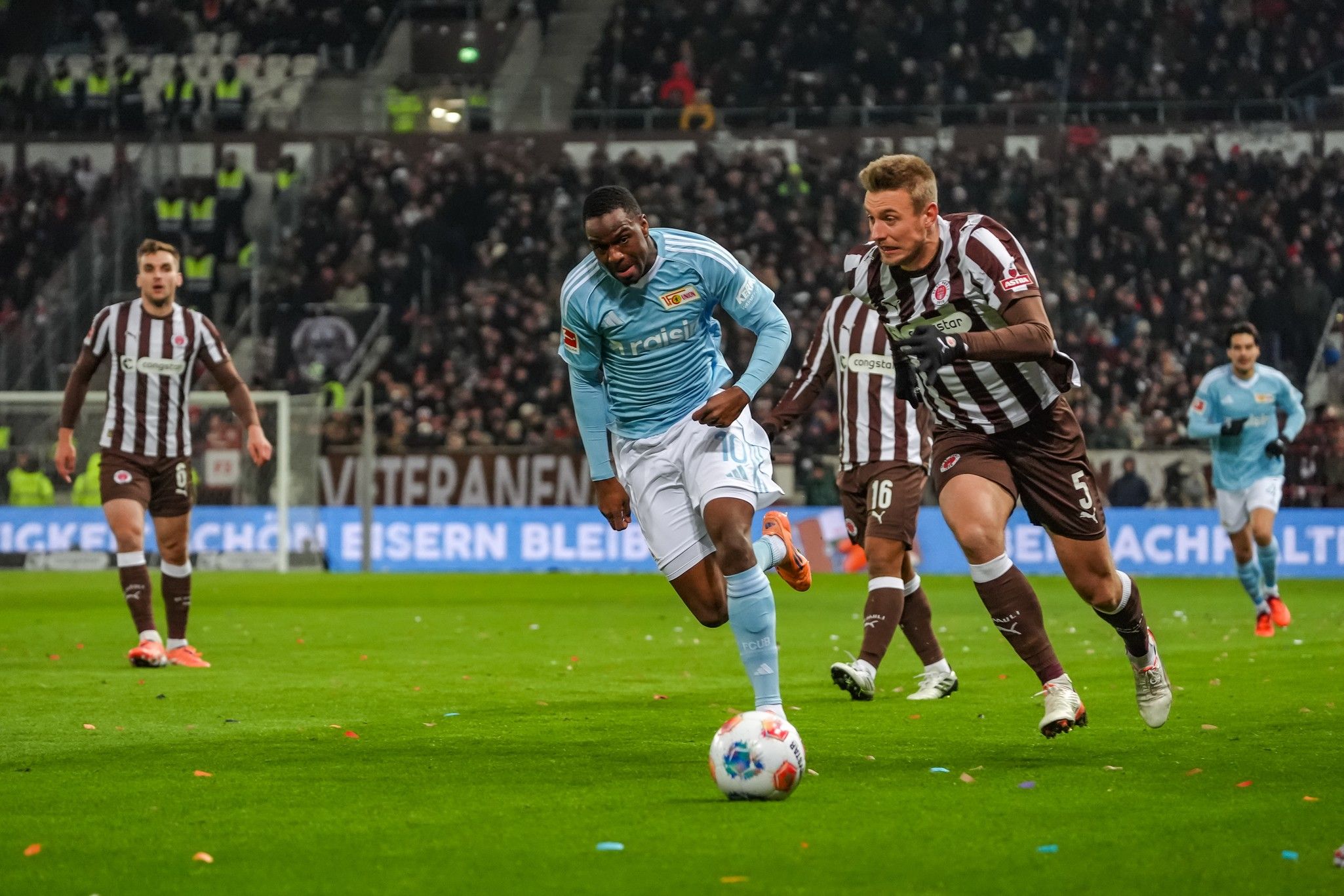 Fußballspielszene mit einem Spieler in hellblauem Trikot, der von einem Gegner in braunem Trikot verfolgt wird.