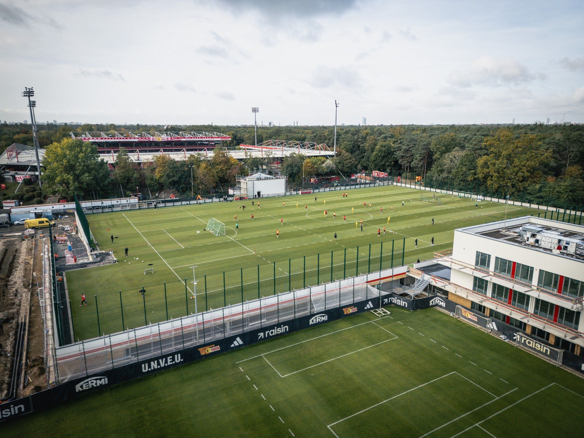 Luftaufnahme eines Fußballfeldes mit Spielern im Training, umgeben von Bäumen und einem Stadion im Hintergrund.