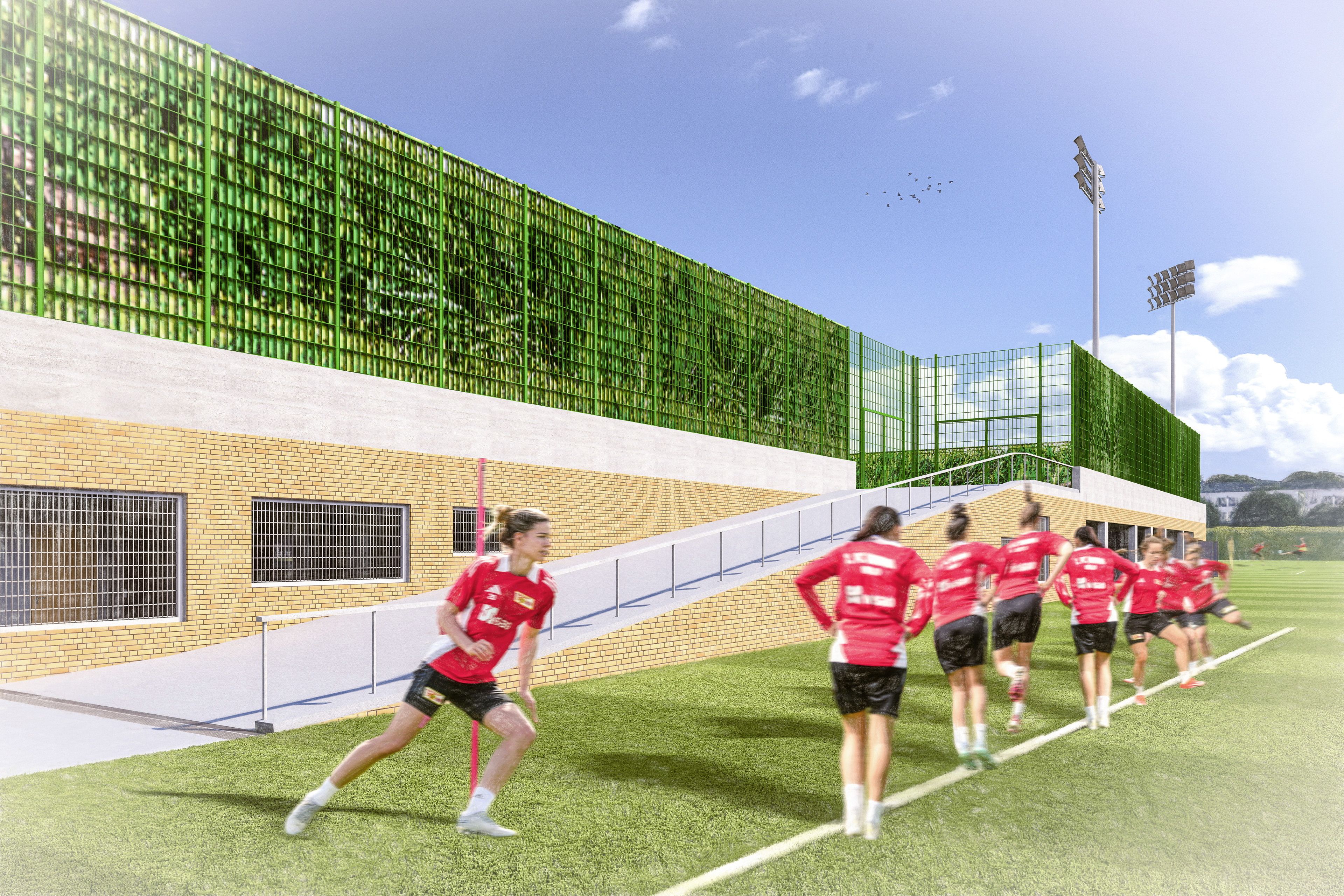 Sportlerinnen in roten Trikots joggen auf einem Fußballfeld, umgeben von einer modernen Trainingsanlage und strahlendem Himmel.