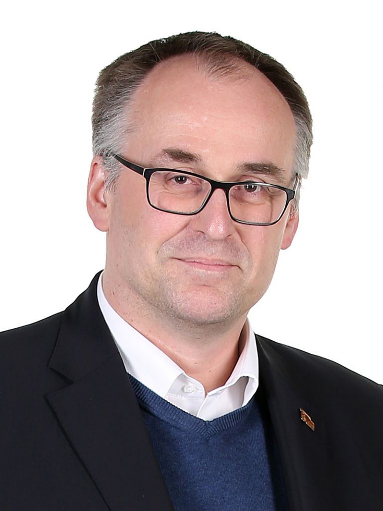 Ein Mann mit Brille, kurzen Haaren und einem leichten Lächeln trägt einen schwarzen Anzug über einem blauen Pullover.