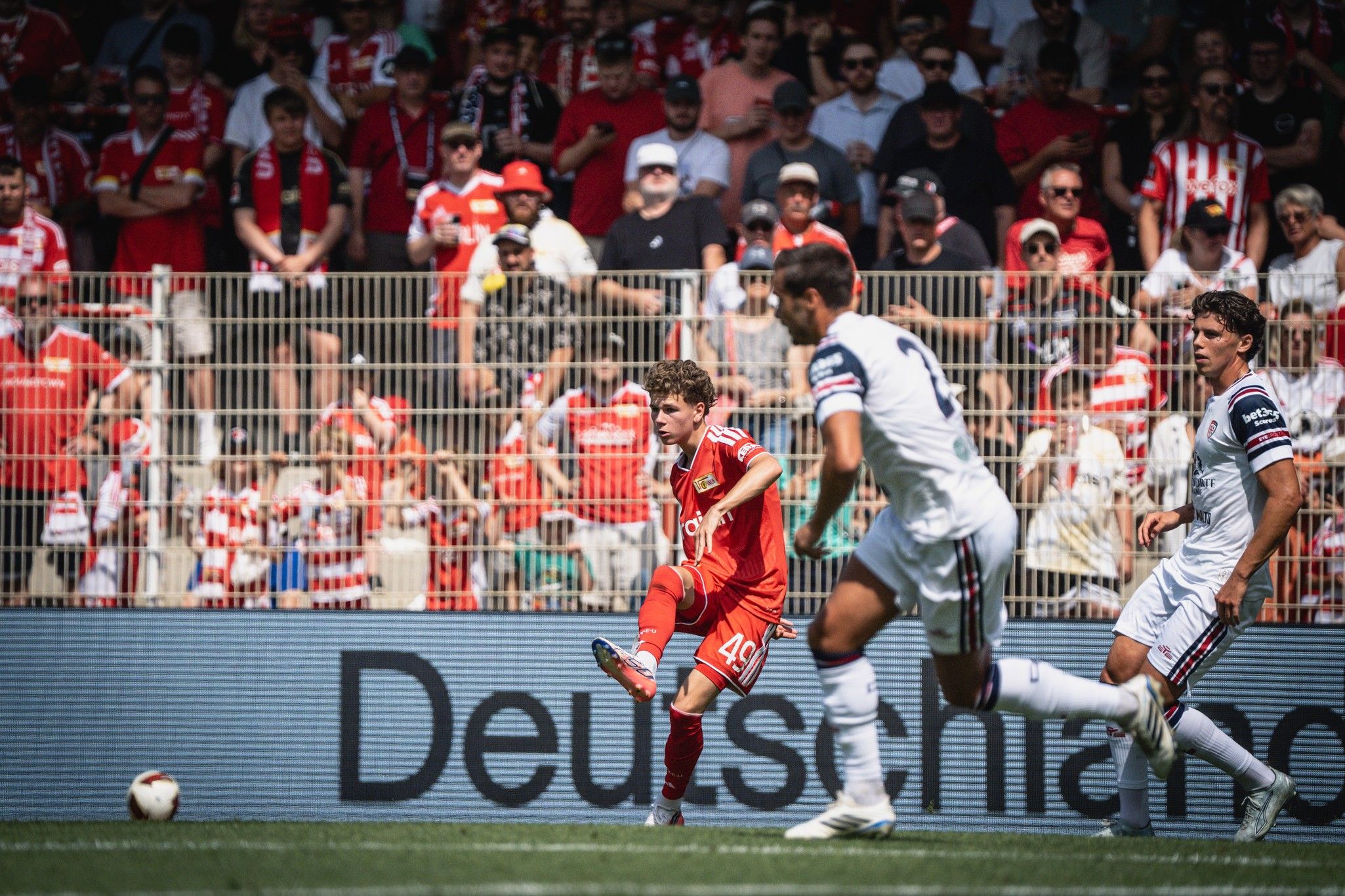 Ein Fußballspieler mit der Nummer 46 schießt auf das Tor, während zwei gegnerische Spieler versuchen, ihn zu blockieren. Fans im Hintergrund.