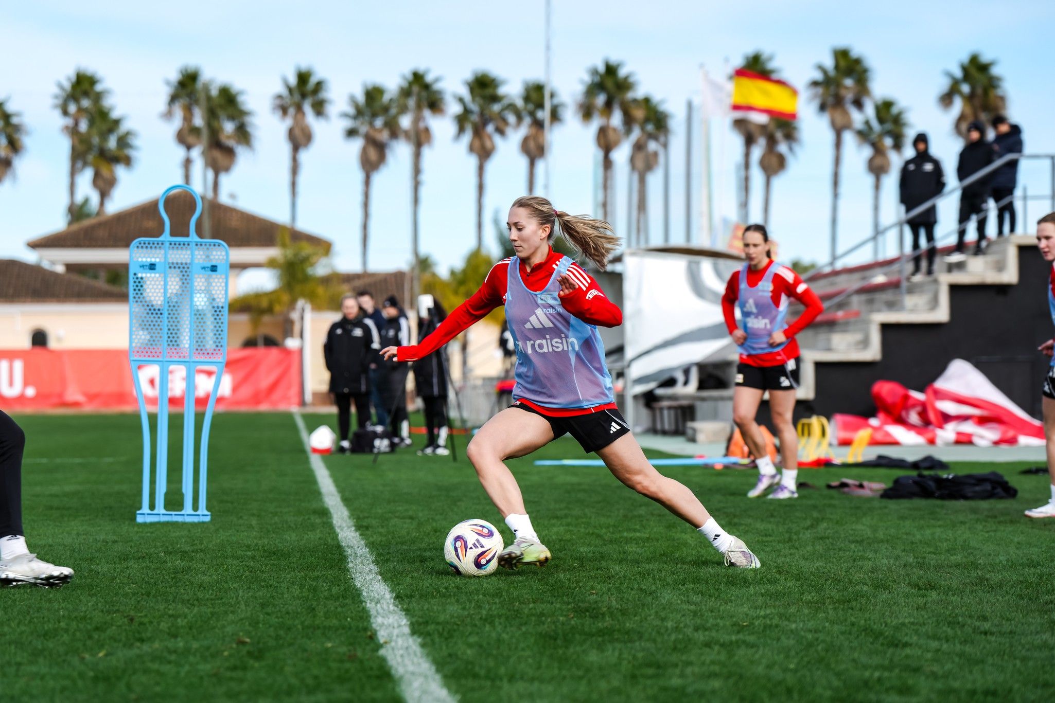 Eine junge Fußballspielerin in Trainingskleidung tritt den Ball auf einem Rasenplatz, umgeben von Palmen und Trainer.
