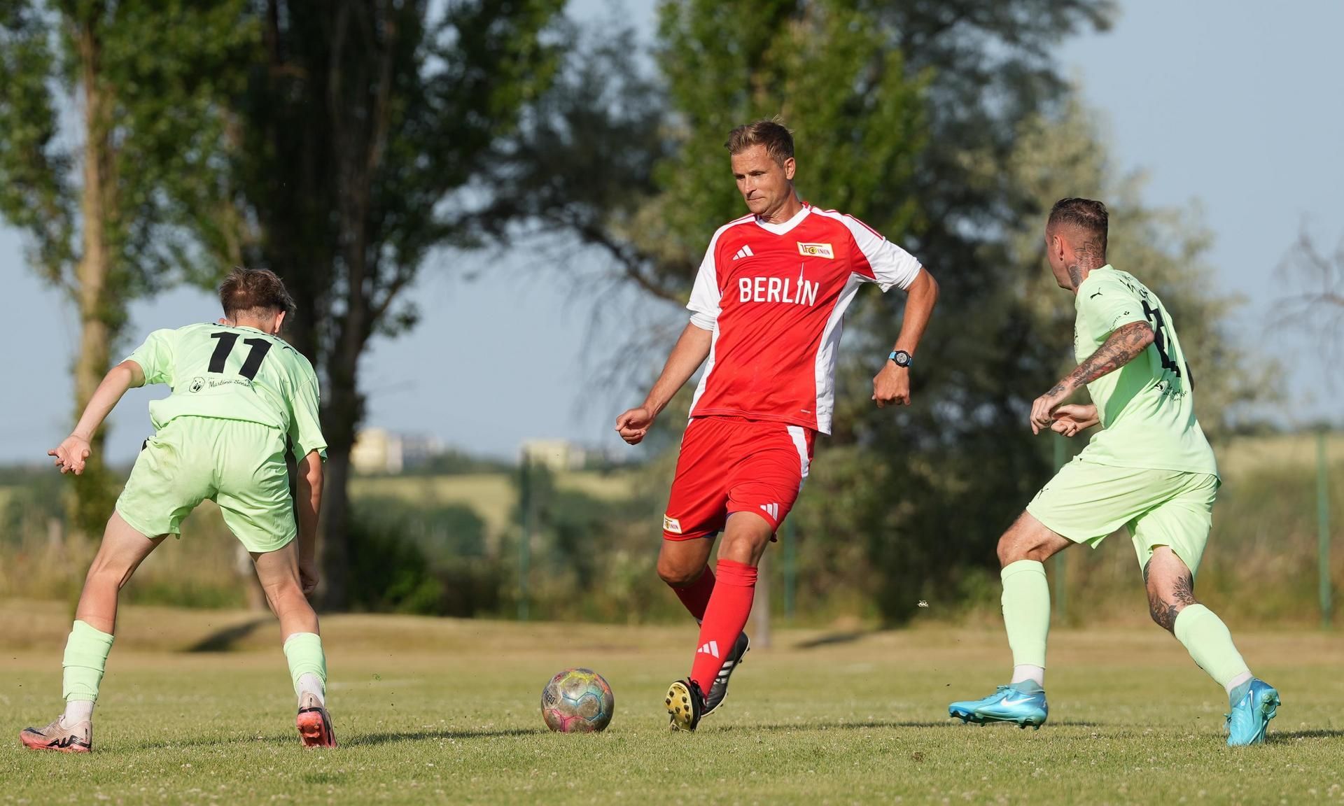 Ein Fußballspieler in rotem Trikot dribbelt den Ball, während zwei Gegner in grünen Trikots ihn anvisieren.
